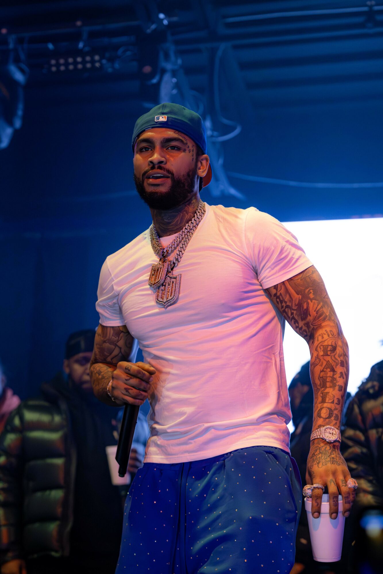 Person with tattoos, wearing a cap and jewelry, holding a microphone on stage with a blue background.