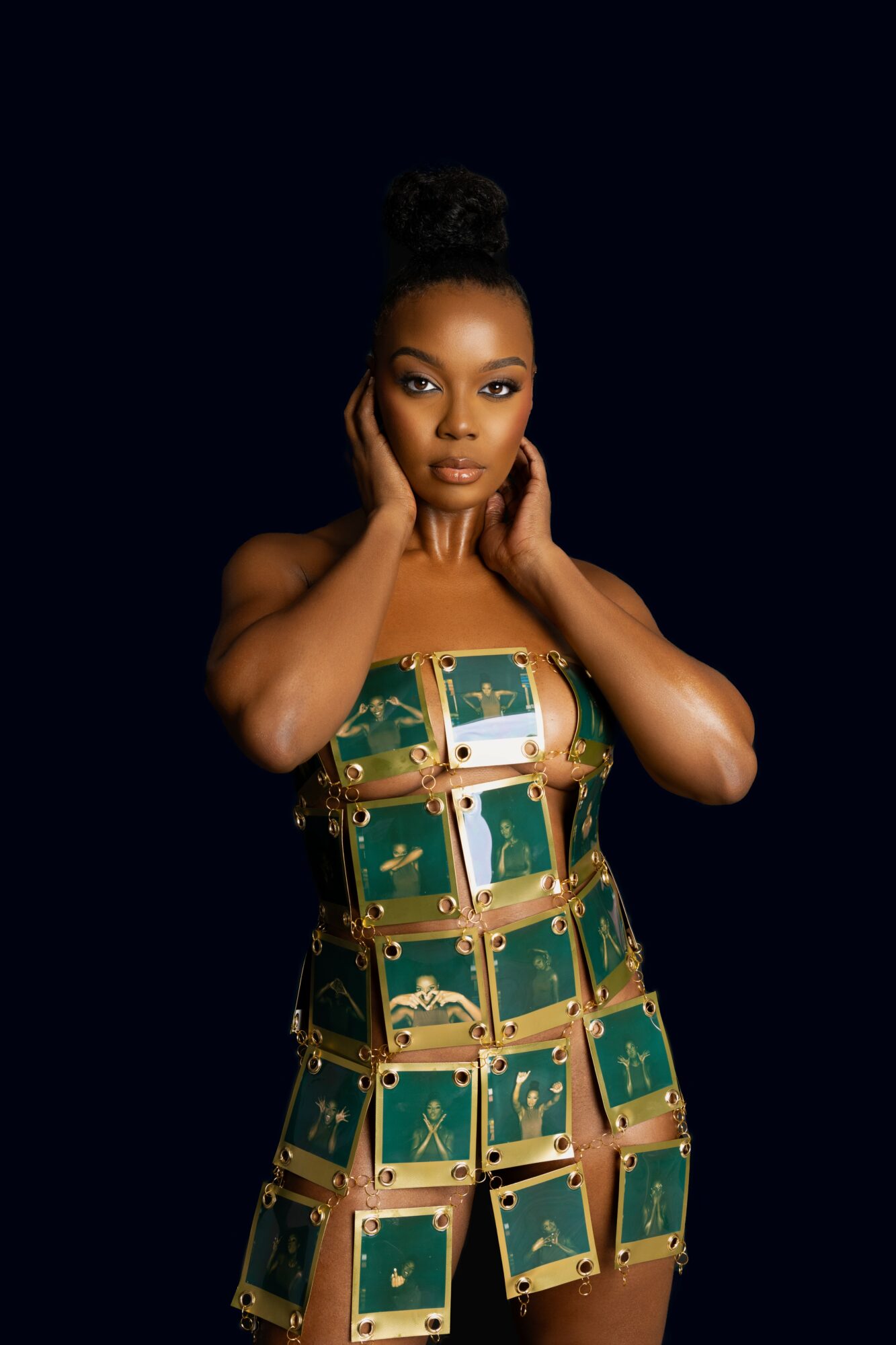 Portrait of a woman with styled hair, wearing a green and gold patterned dress, posing with hands near face against dark background.