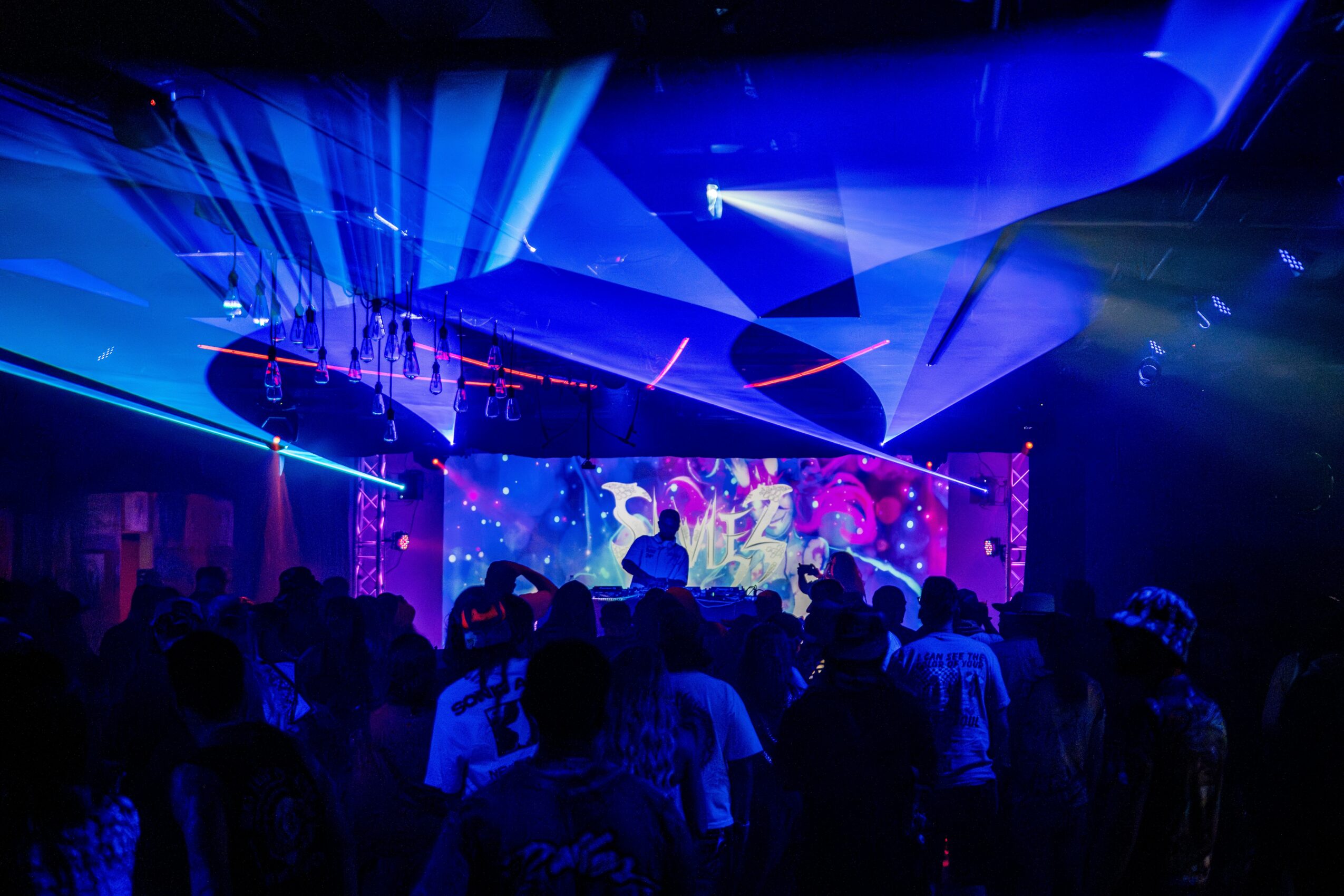 Crowd watching a colorful stage with bright lights and laser effects in a nightclub or concert venue.