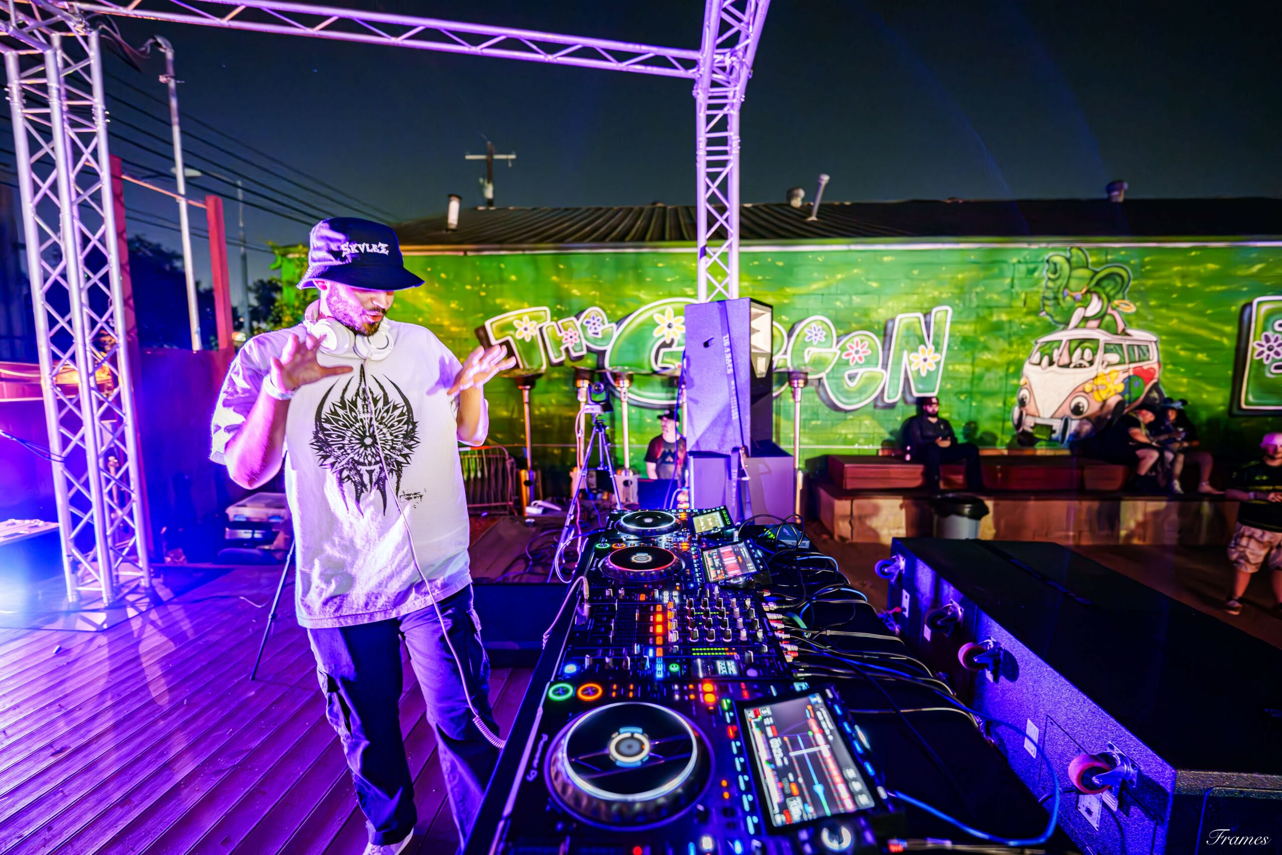 DJ wearing a cap and t-shirt, standing at a DJ booth with equipment, stage with lights, and a large screen in the background.