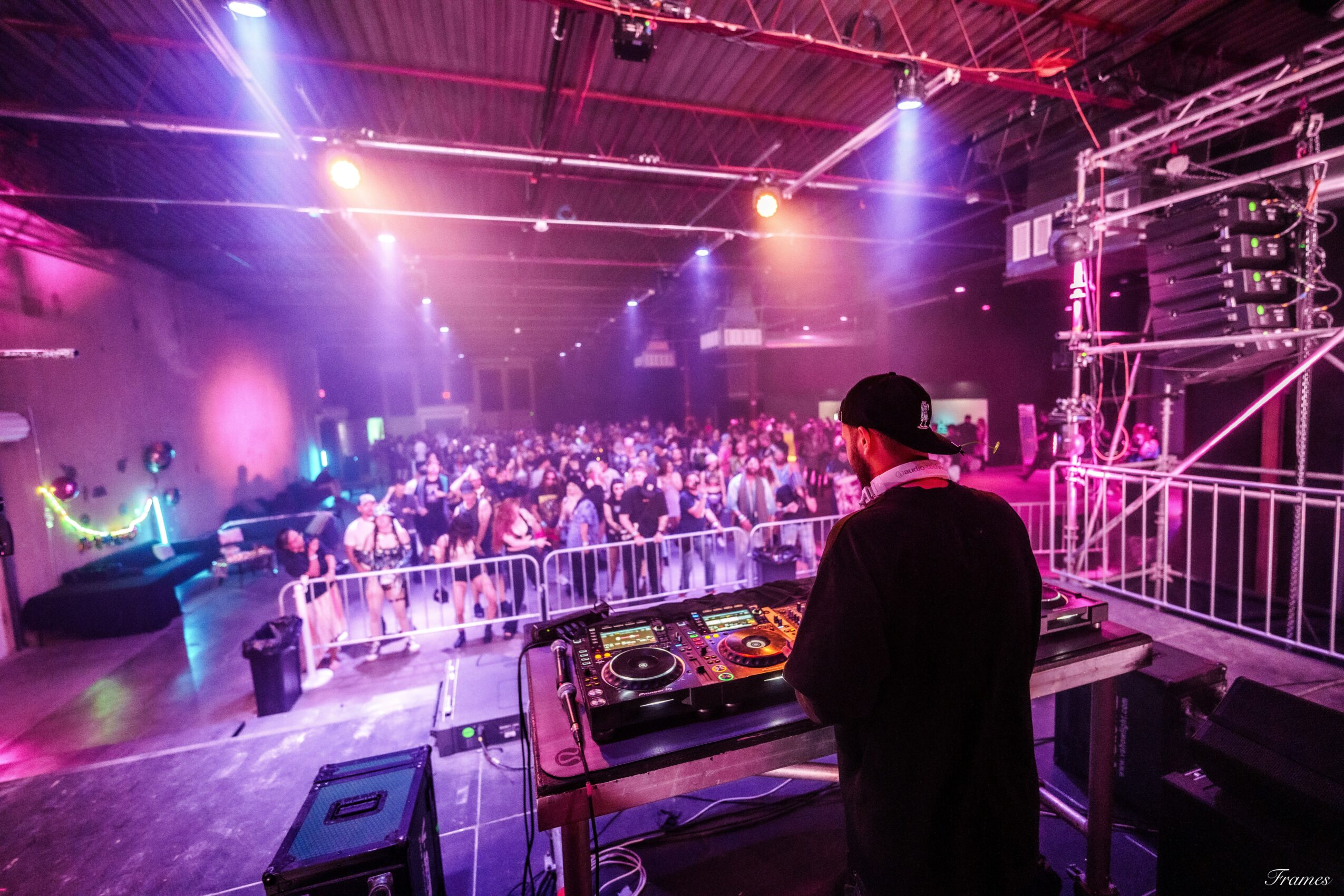 DJ in black attire and cap performs at a crowded indoor dance event with colorful lighting and a large audience.