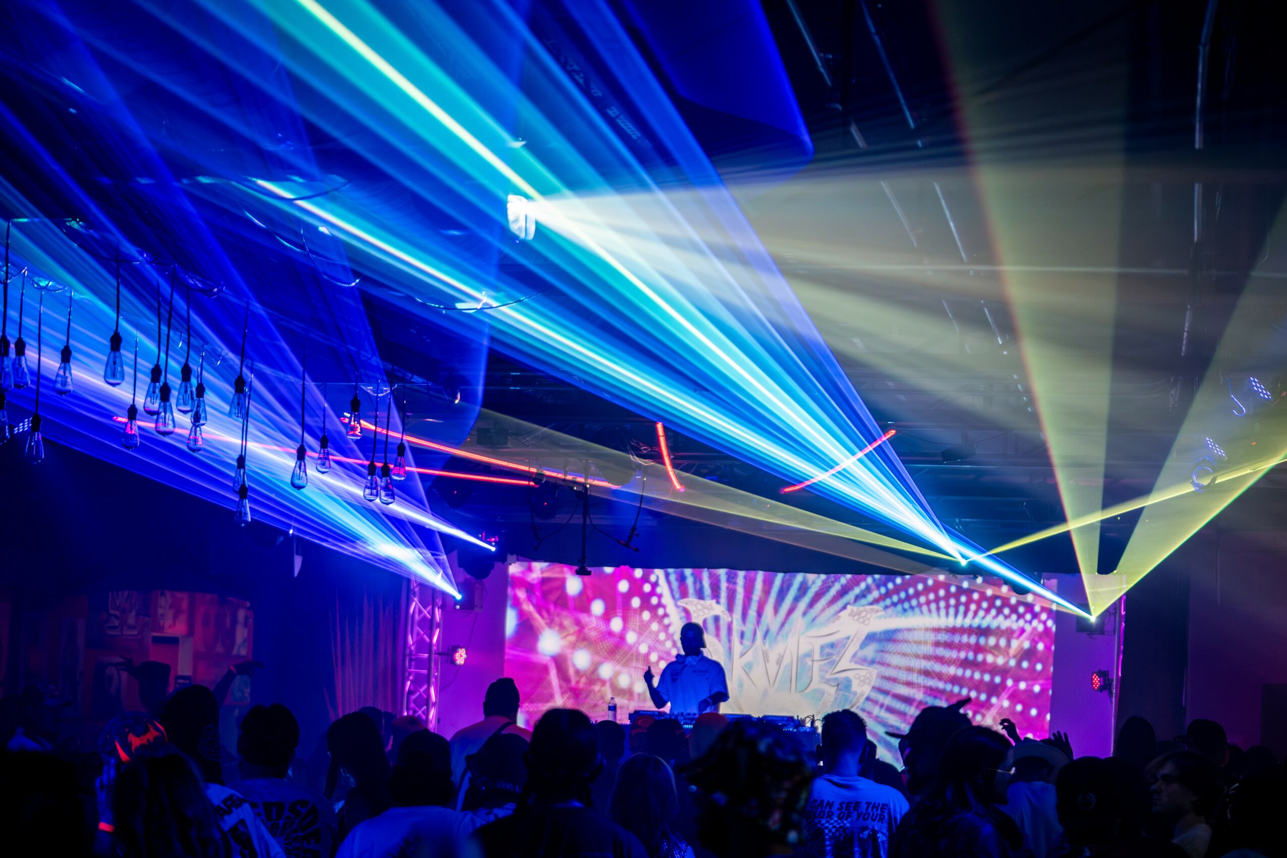Concert scene with colorful laser lights, a DJ on stage, and an audience in a dark room.