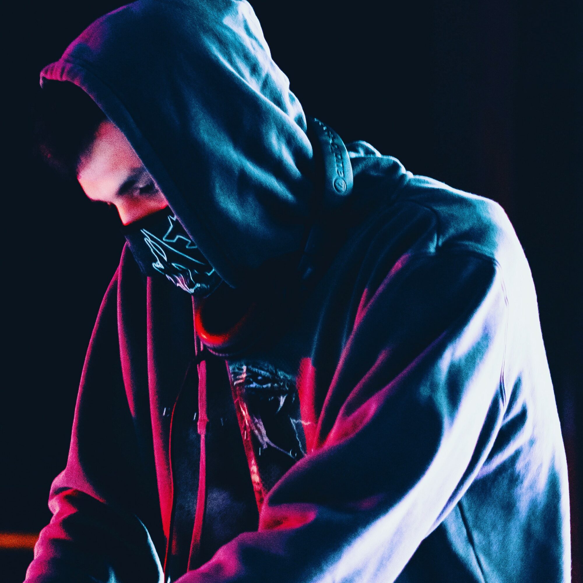 Person wearing a hoodie and face mask, looking down, with neon lighting effects, dark background.