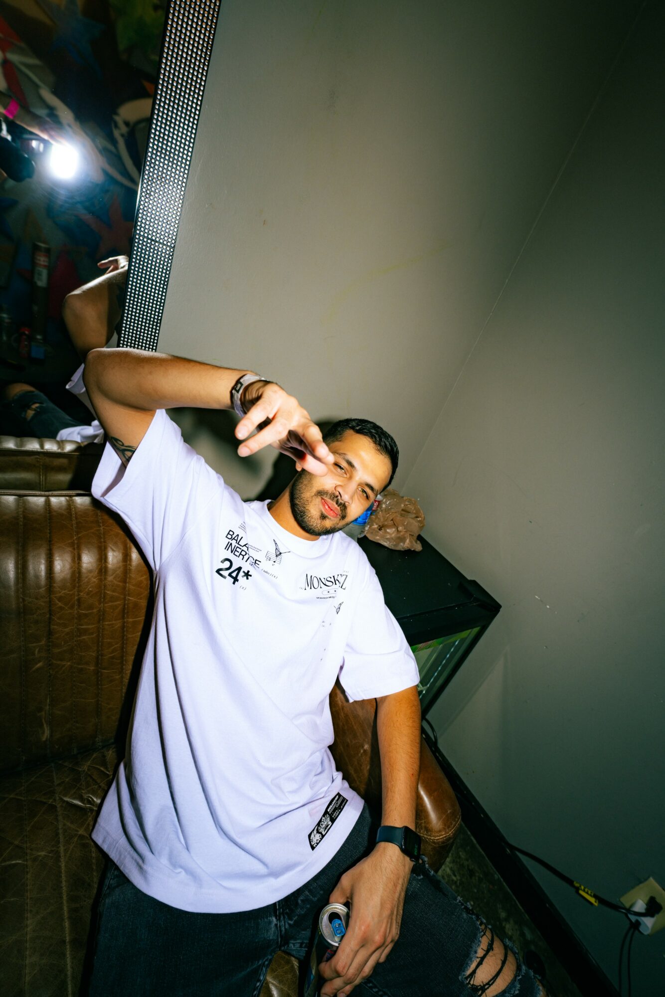 Man with a beard and short hair, wearing a white t-shirt, smiling and holding a drink, sitting indoors.