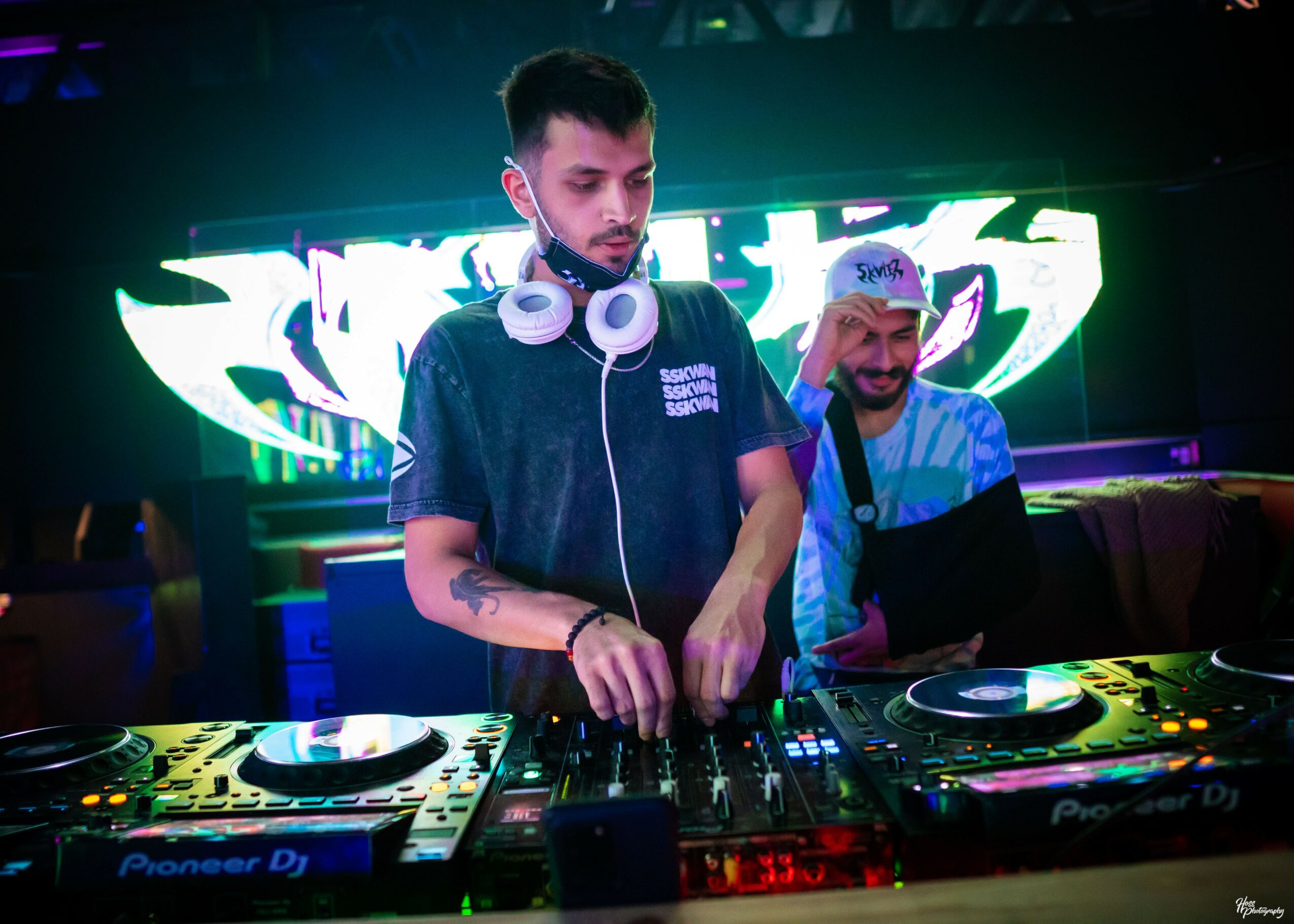 DJ with headphones around neck, mixing music at a club with colorful background lights.