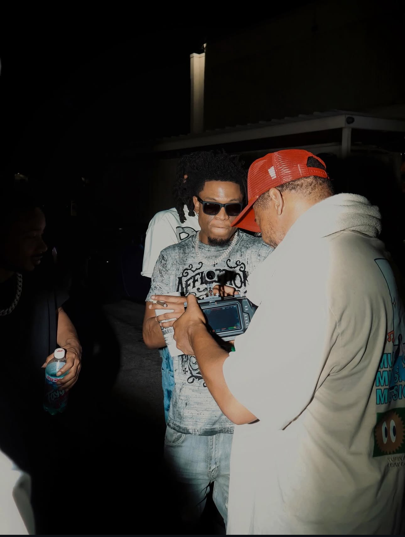 Two men are looking at a camera, one wearing sunglasses and a graphic t-shirt, the other in a hoodie and red cap.
