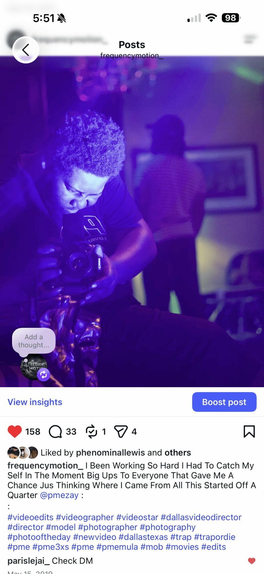Person with curly hair looking at a camera, with a blurred background of another person and framed picture, purple lighting