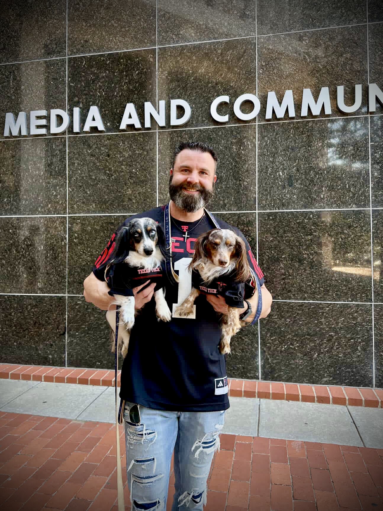Man holding two dogs in front of a wall with partially visible text, smiling, wearing a dark jacket and ripped jeans.