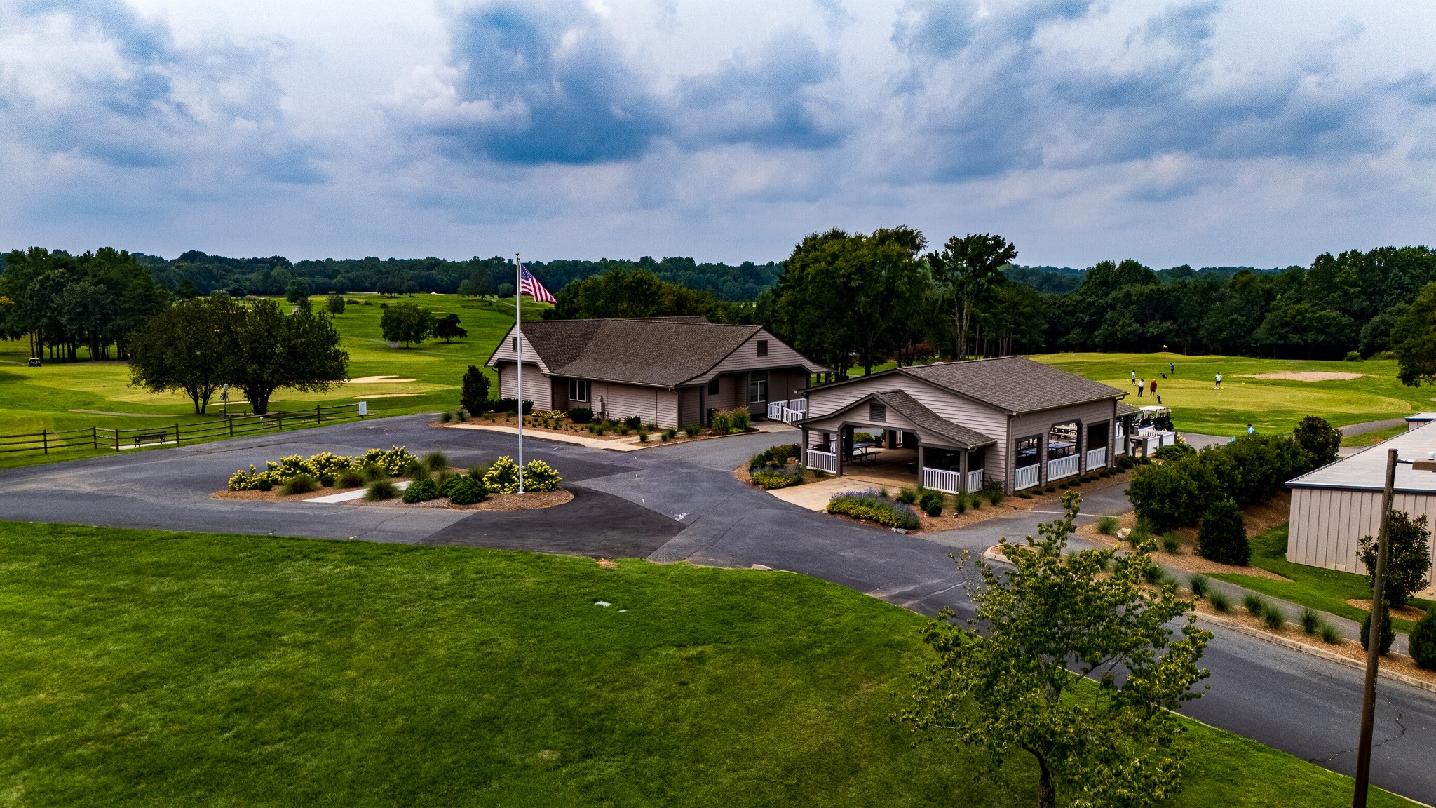 A building with a parking lot, surrounded by green grass and trees, under a partly cloudy sky.