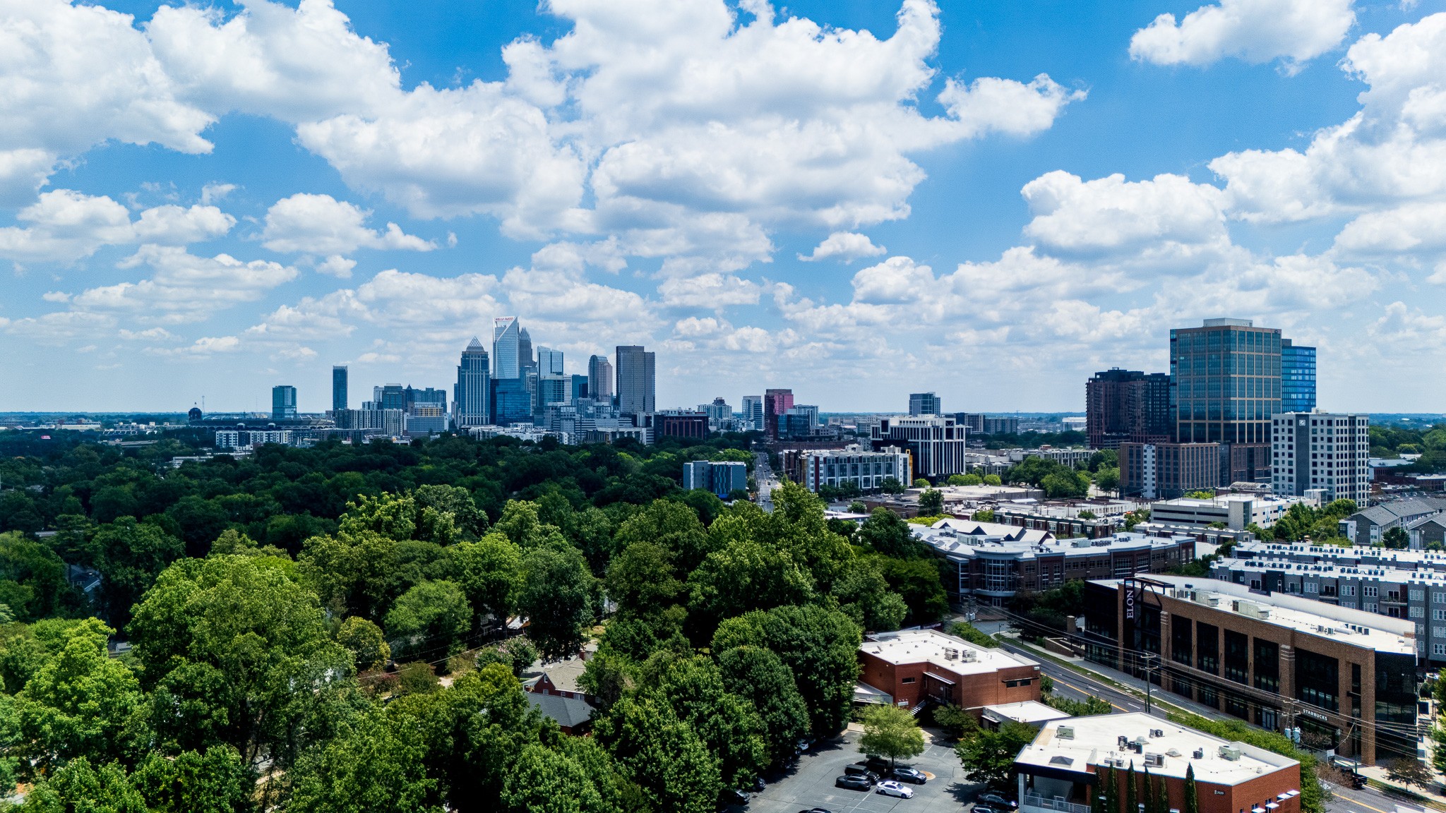 City skyline with tall buildings, surrounded by green trees and a partly cloudy sky.