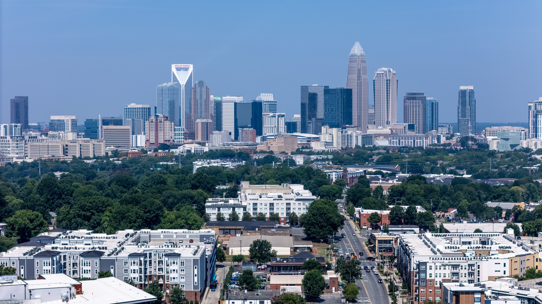 City skyline with tall buildings, surrounded by greenery and residential areas, under a clear blue sky.