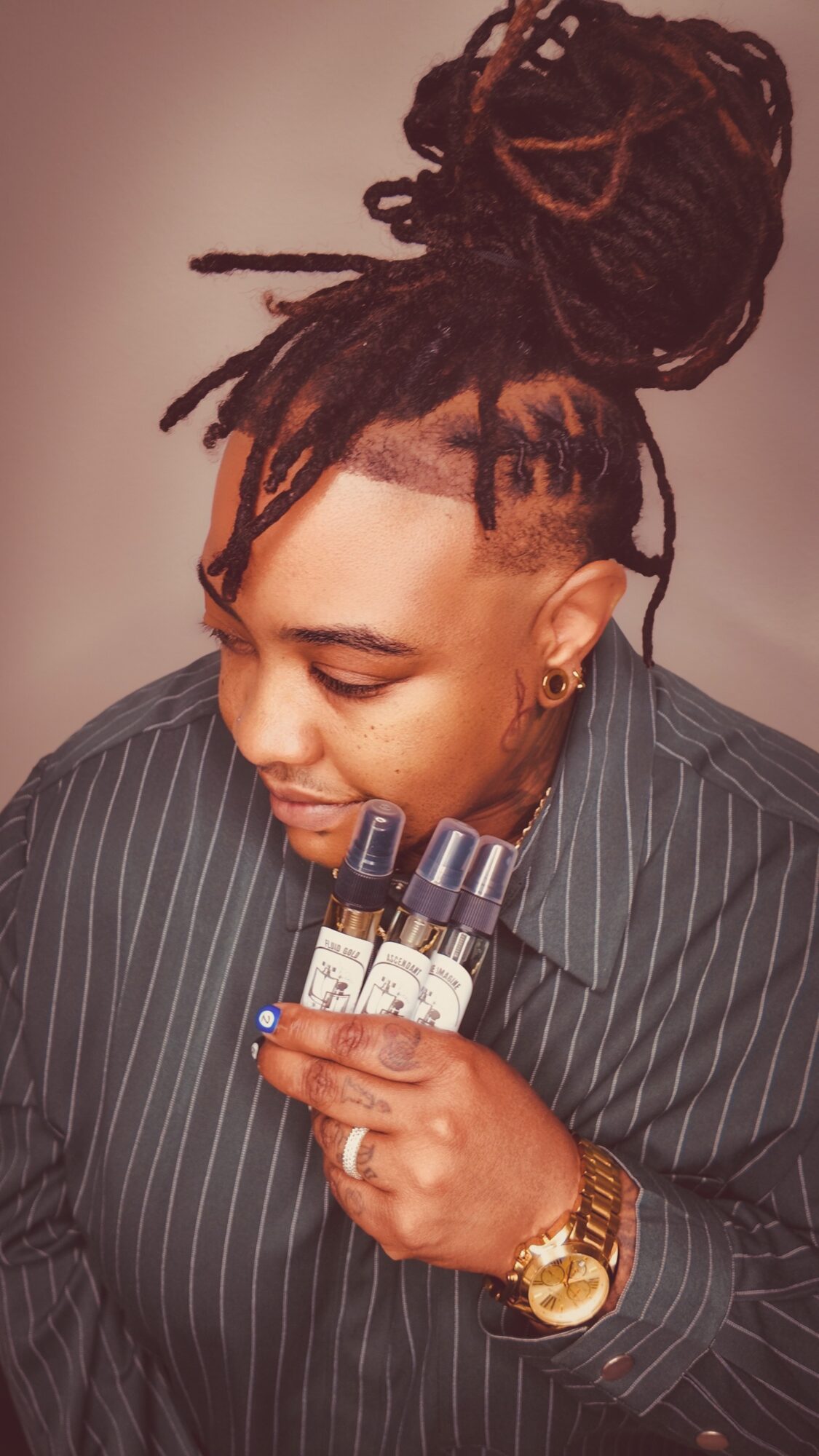 Person with dreadlocks styled in a bun, wearing a pinstripe shirt, holding perfume bottles, and wearing a watch and earrings.