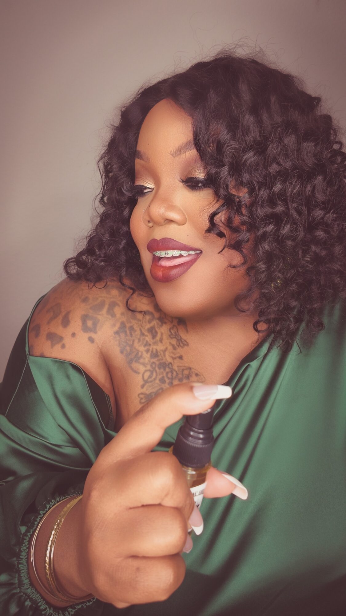 Woman with curly hair holding a small spray bottle, smiling, wearing a green satin top, with tattoos on her shoulder.
