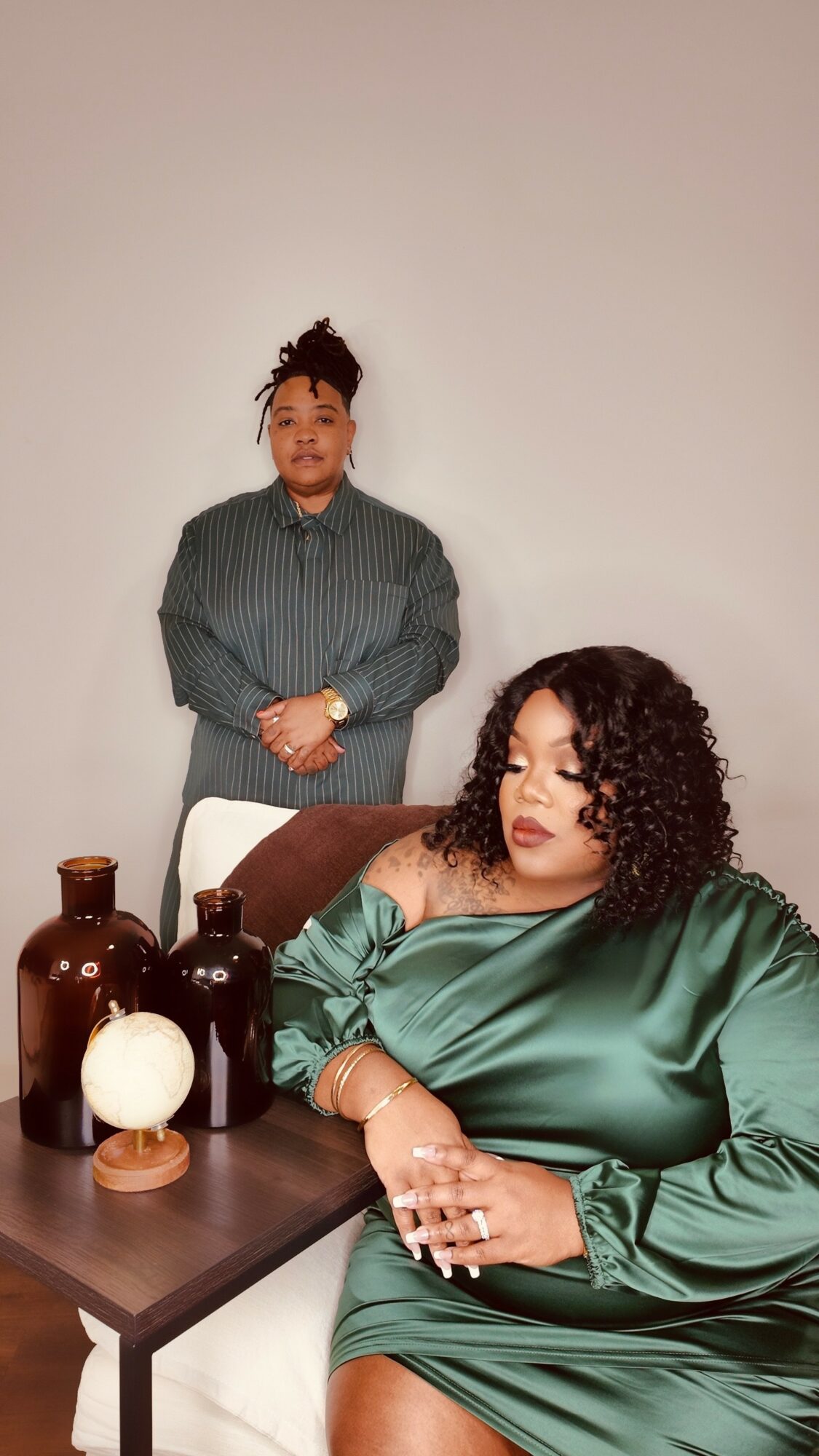 Two women in a room with a plain background, one sitting at a table and the other standing behind her.