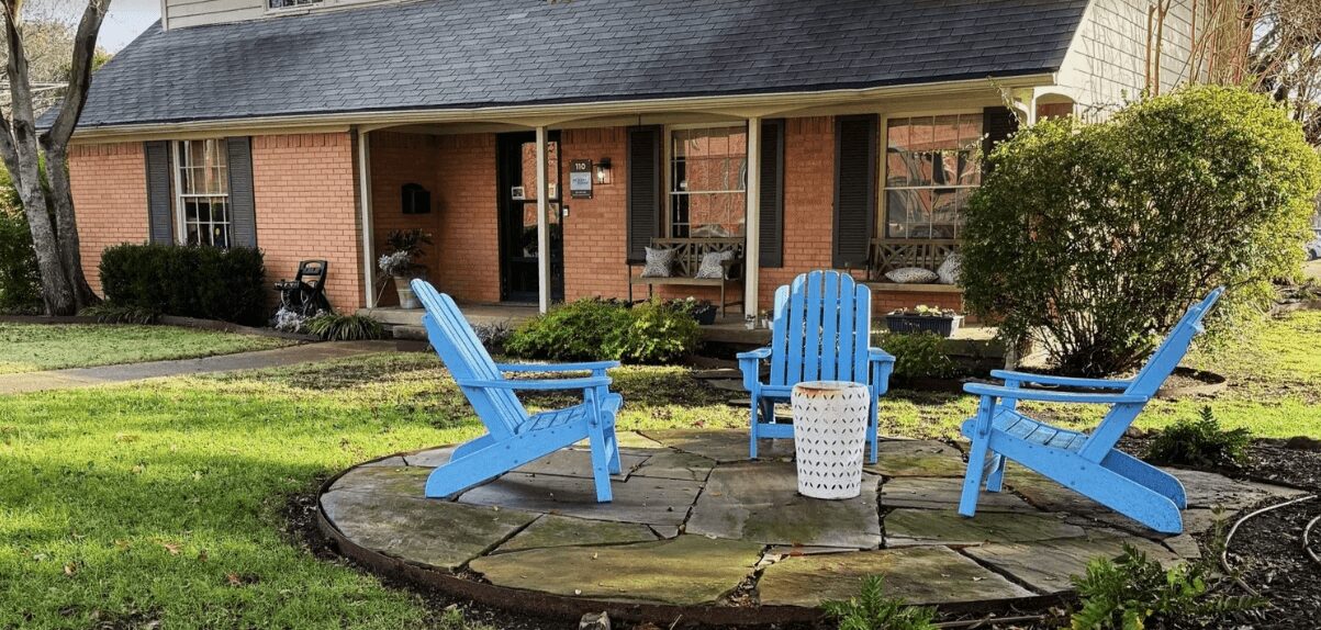 Three blue Adirondack chairs around a small white table on a circular stone patio in a yard with grass and bushes.
