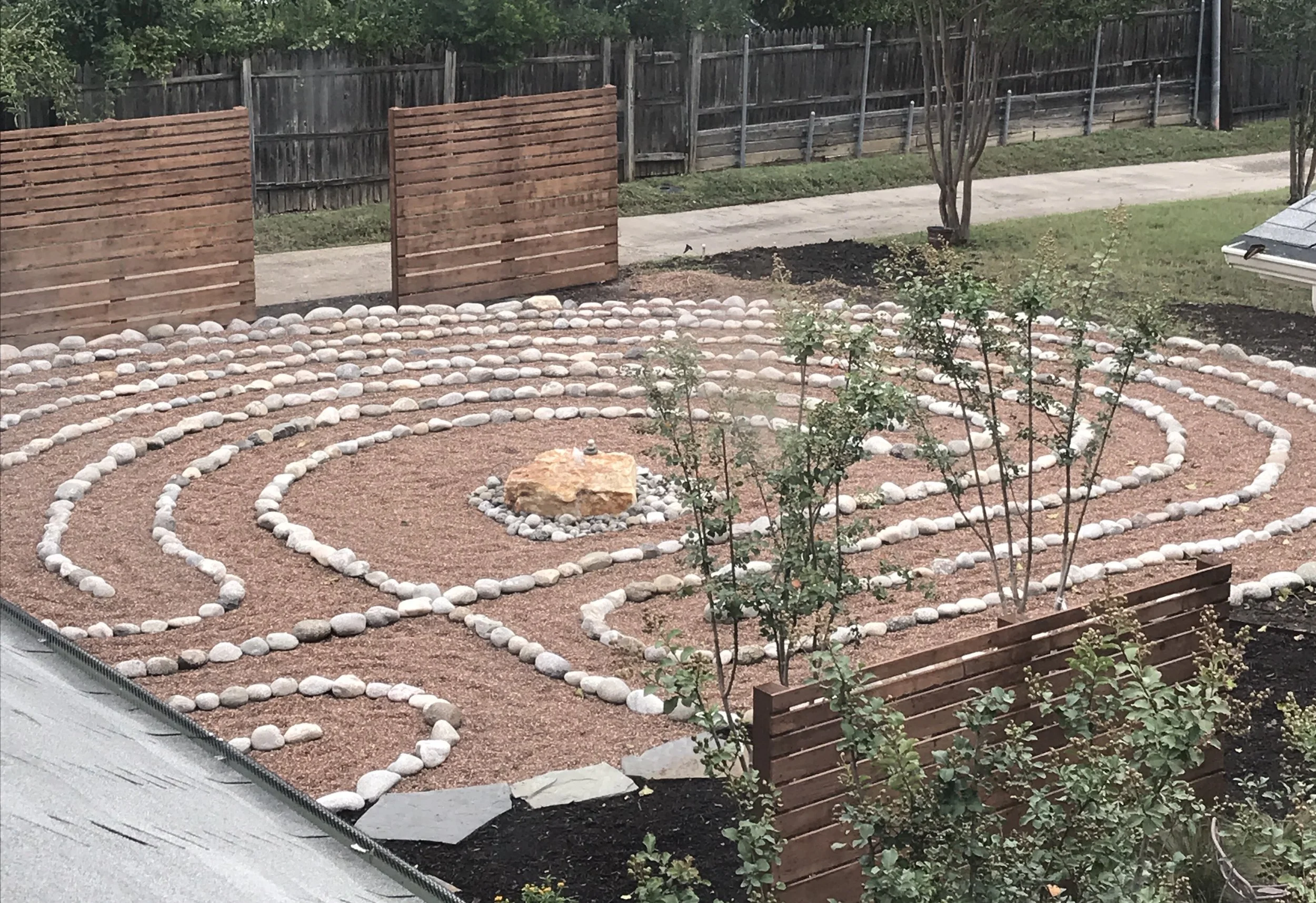 Decorative garden with circular stone pathways, small plants, and a central feature, enclosed by a wooden fence.