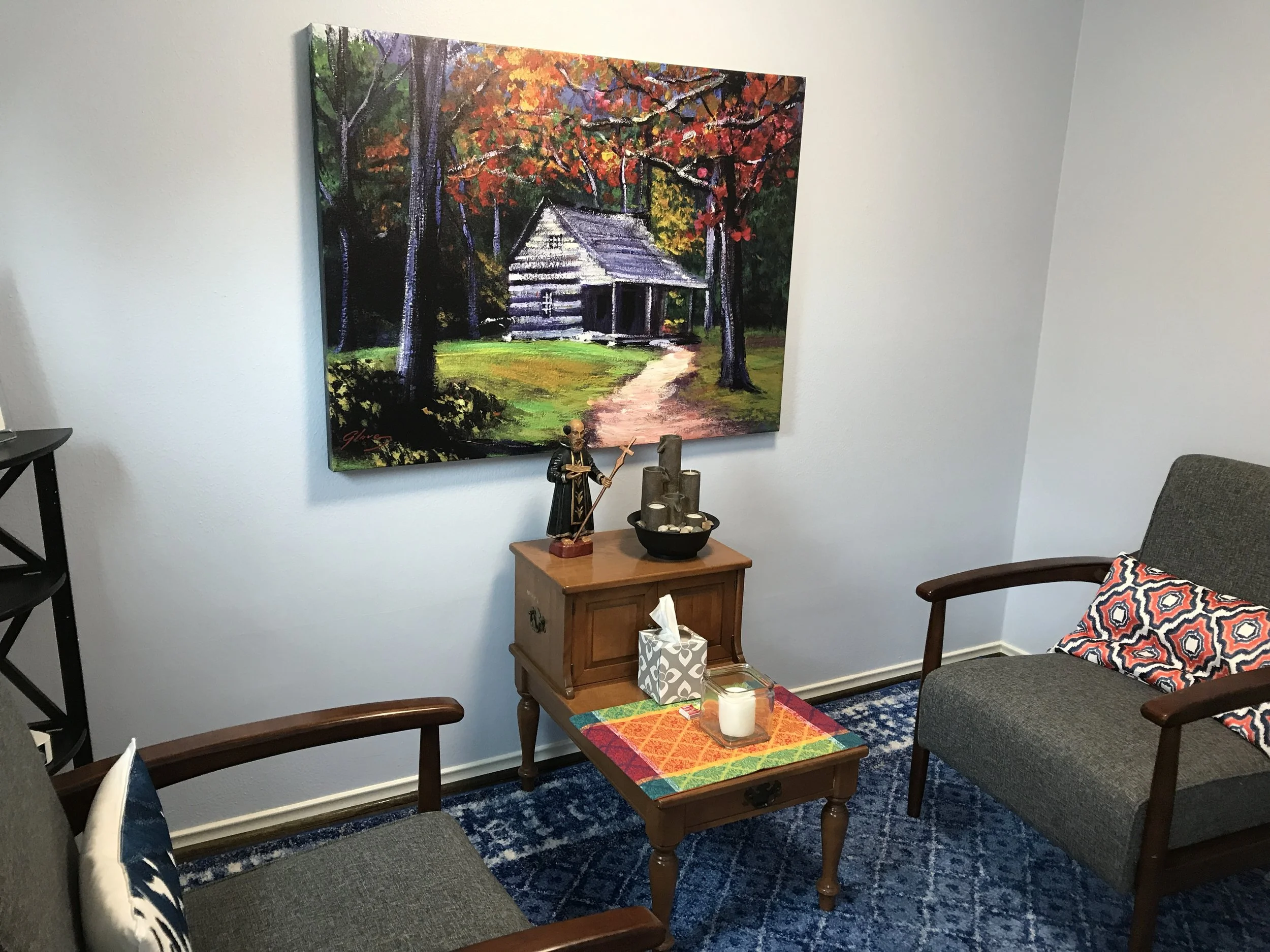 Painting of a house surrounded by trees with colorful leaves, hanging on a white wall in a room with chairs and a small table.