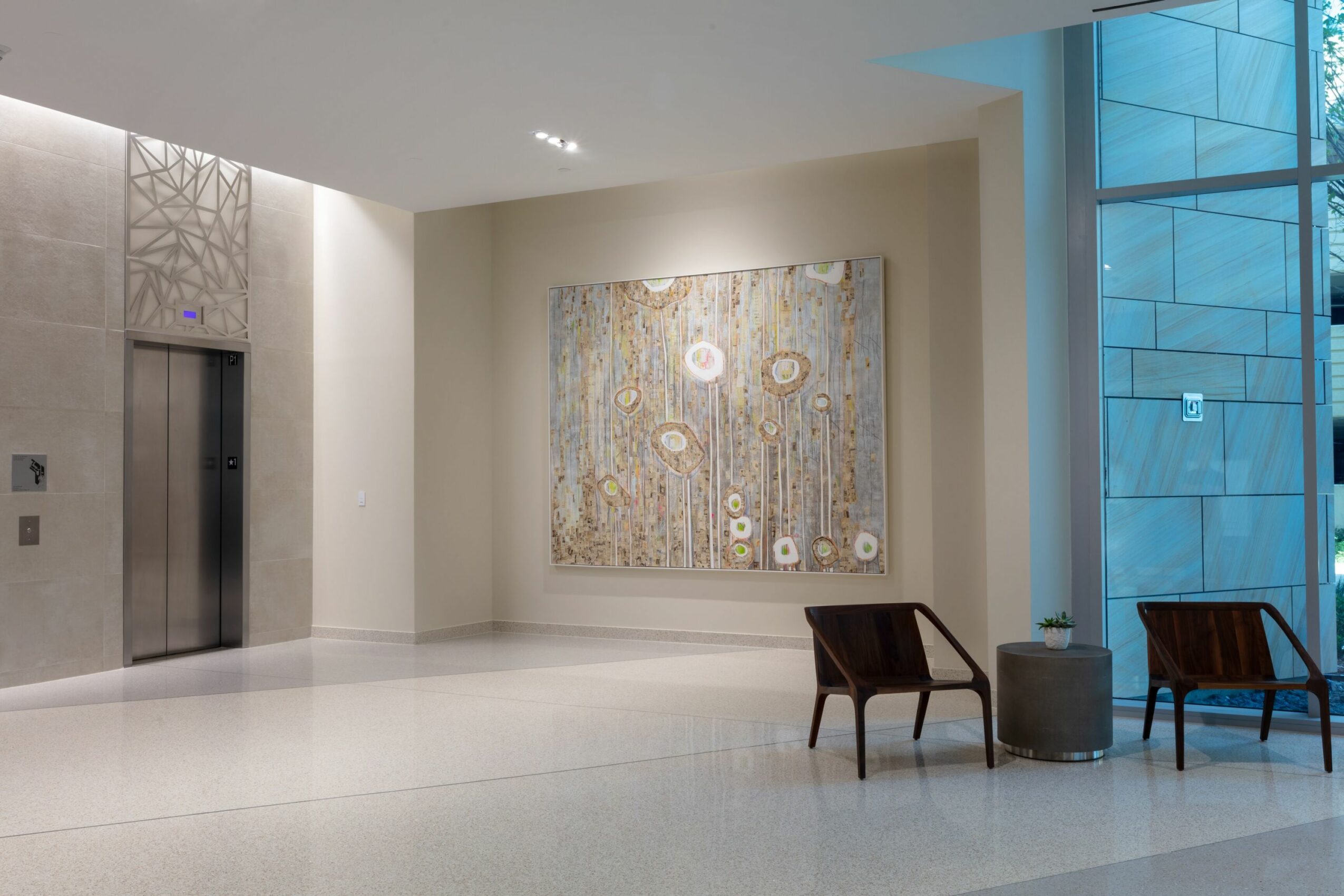Lobby with elevator, abstract painting on wall, two chairs, and large blue-tinted window, bright and modern interior.
