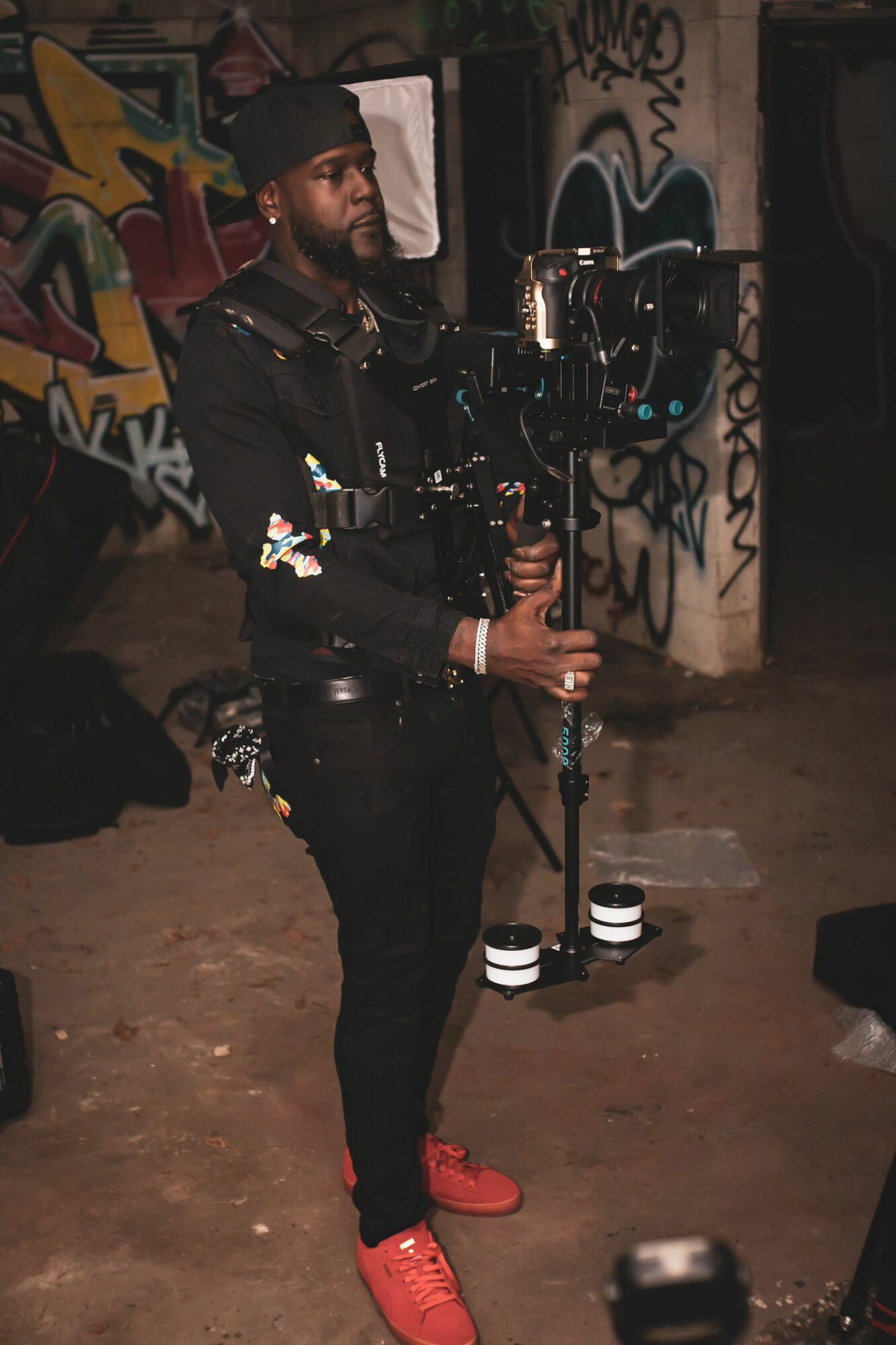 Person holding camera stabilizer in front of graffiti-covered wall at night, wearing black clothing and red shoes.