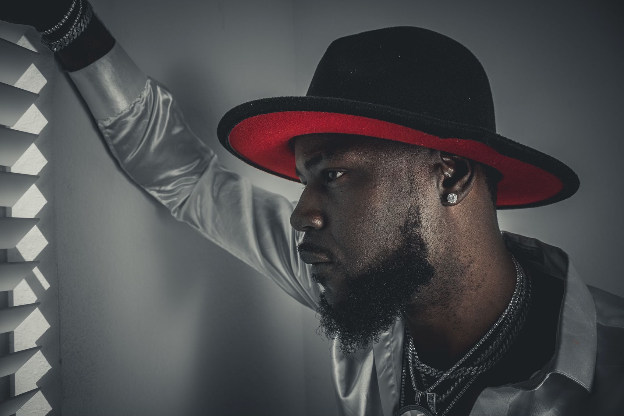 Man wearing a black hat with red band, silver shirt, and jewelry, looking to the side, with a neutral background.