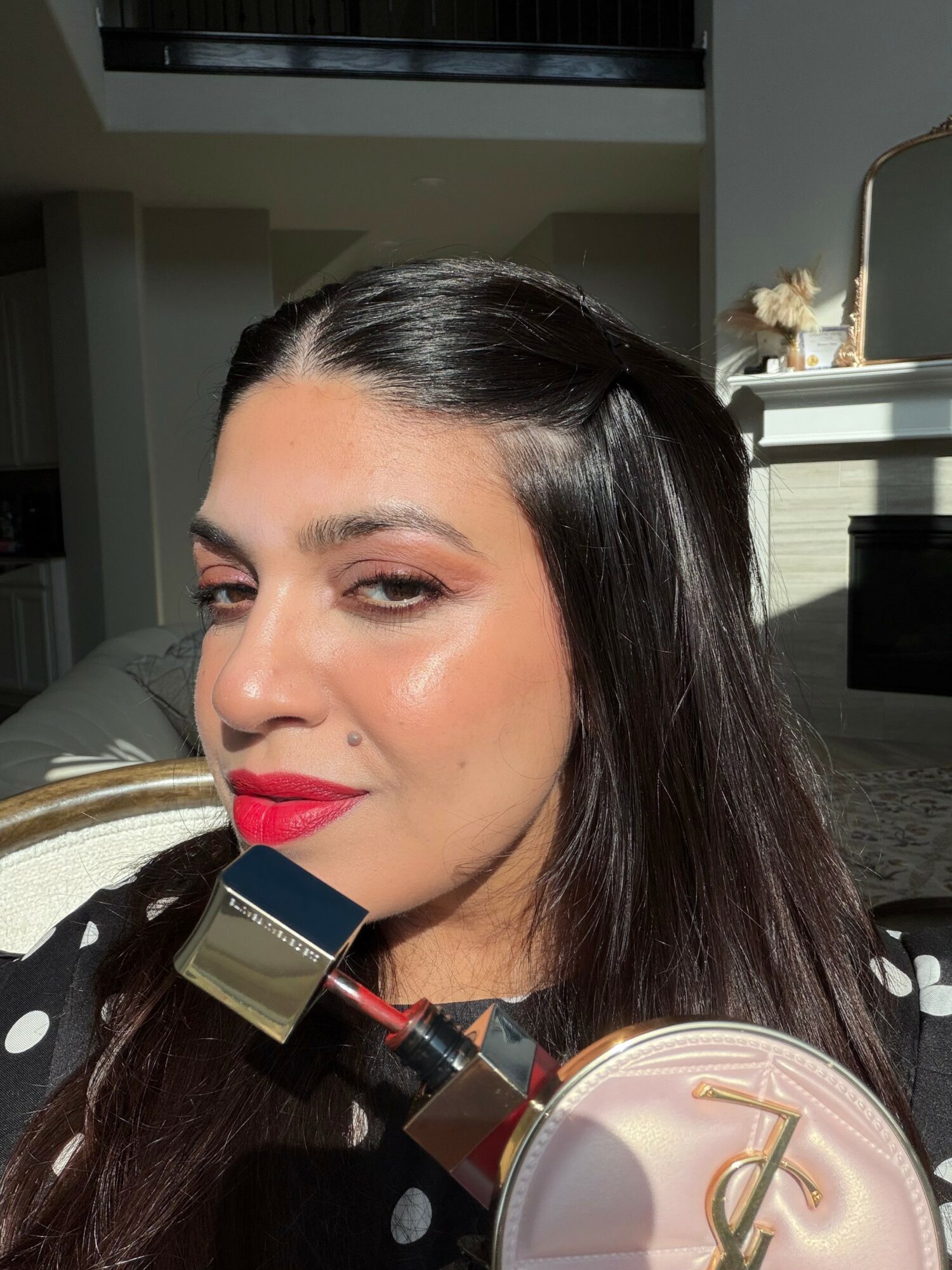 Close-up of a woman with dark hair, red lipstick, and makeup, holding a makeup brush near her face, in a well-lit room.