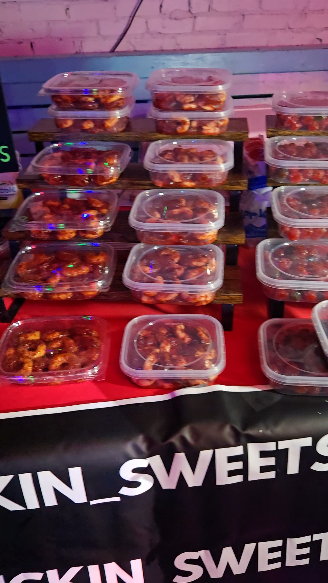 Multiple clear plastic containers filled with colorful candies arranged on a table with a black and red cloth, pink brick wall background.