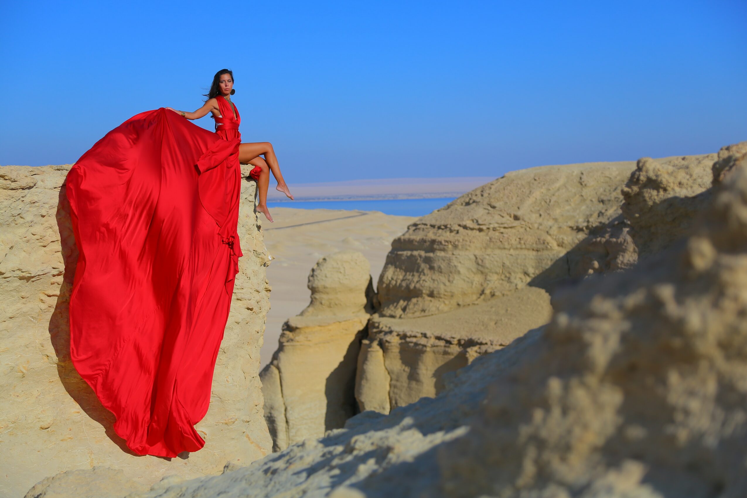 Woman in a red dress sitting on a rock formation in a desert landscape with blue sky.