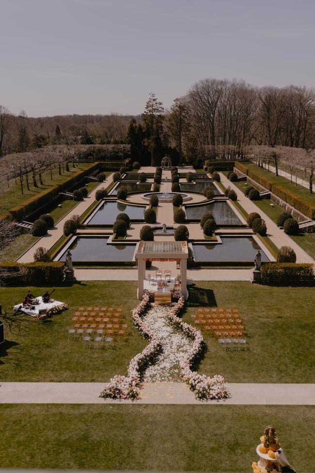 Formal garden with symmetrical water features, hedges, and a central pathway, viewed from above. A sculpture of a dog with flowers is in the foreground.