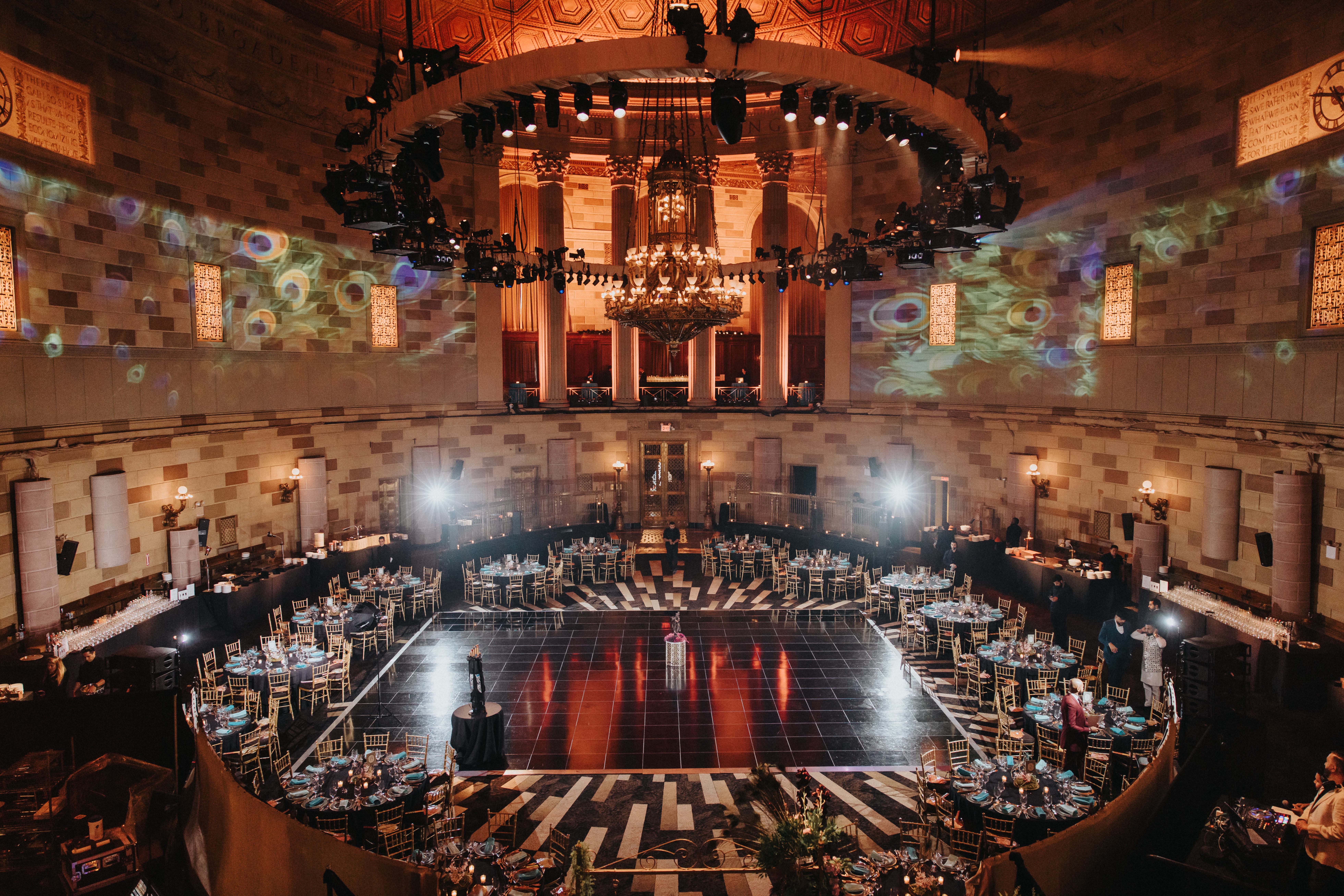 Large banquet hall with a central dance floor, round tables, and a chandelier hanging from the ceiling, illuminated with colorful lighting.