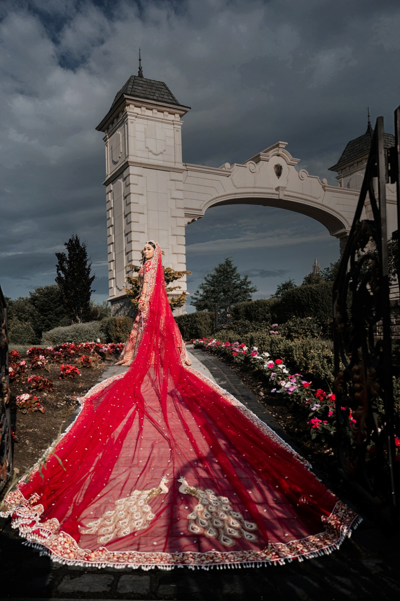 Person in a red dress with a long train standing in front of an ornate archway and gate, garden with flowers, cloudy sky