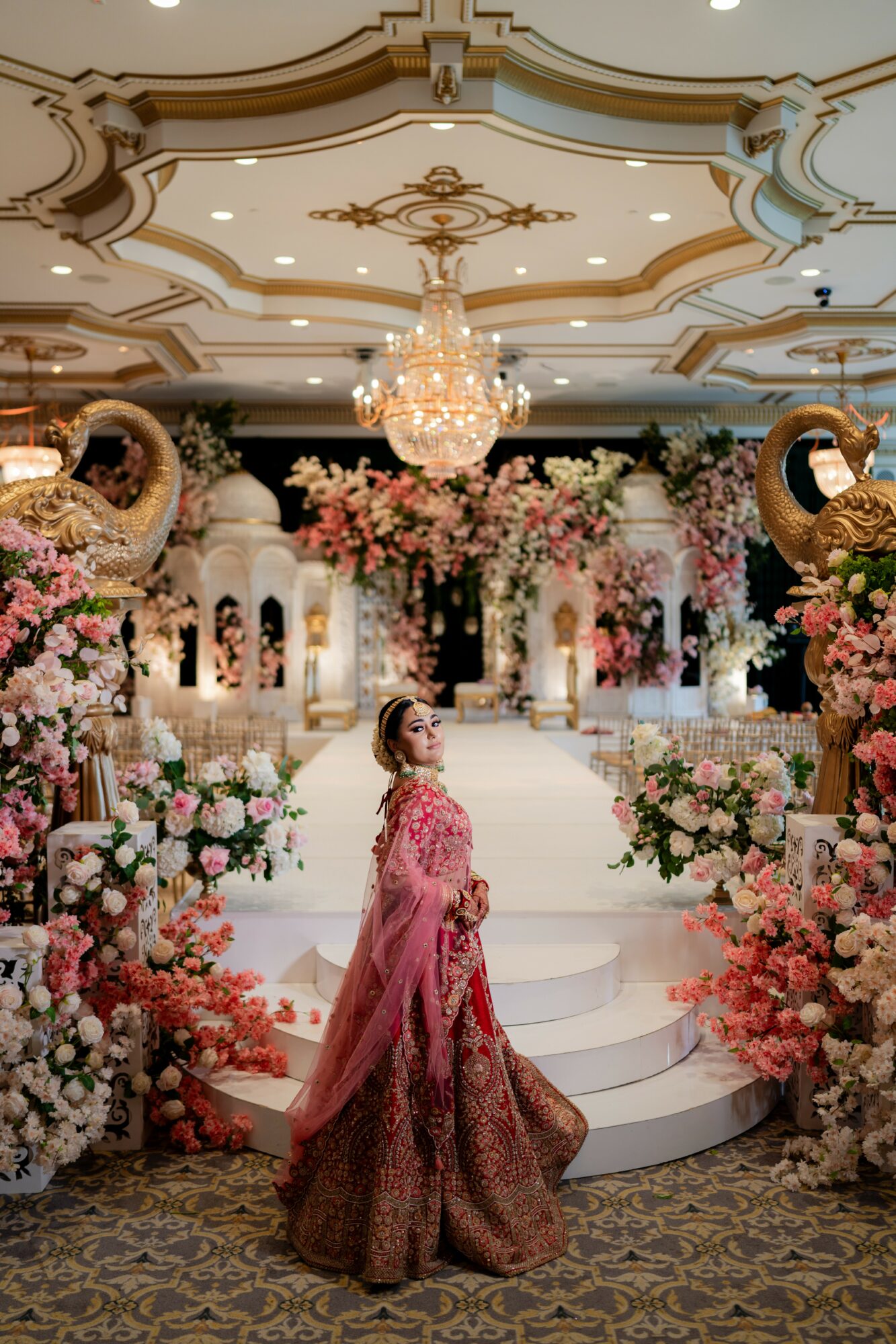 A woman in a pink and gold gown stands on a decorated stage with floral arrangements and chandeliers.