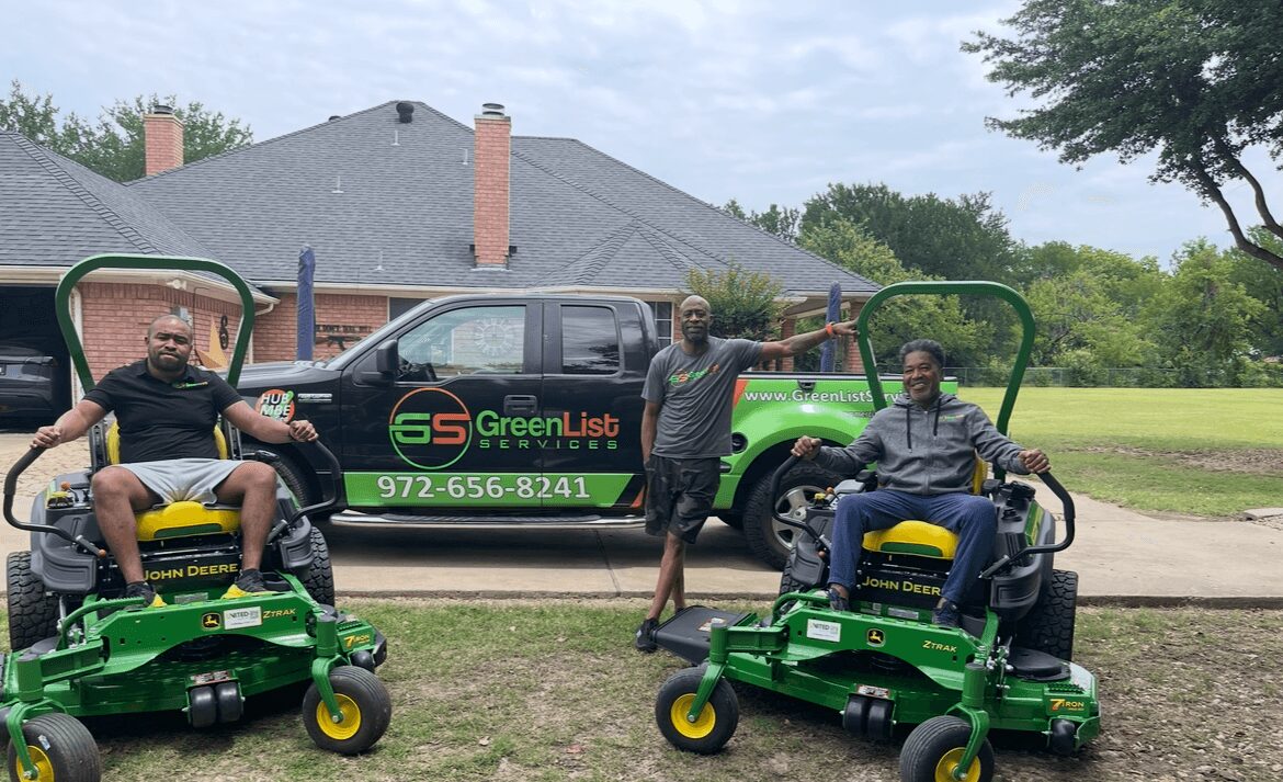 Three people outdoors with two riding lawn mowers and a truck in the background, trees, and a house. Two men sit on mowers, one stands between them.