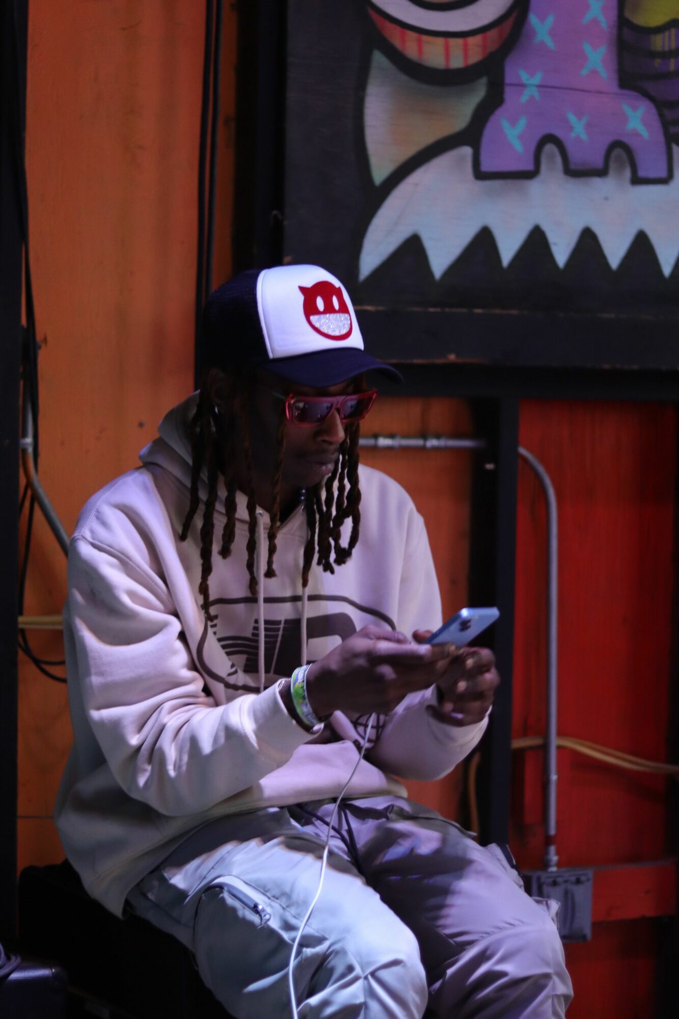 Person with dreadlocks wearing a white hoodie and black cap, sitting and using a phone, with colorful wall art behind.