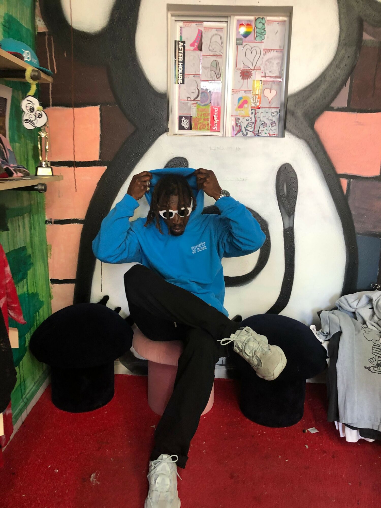 Person with dreadlocks in a blue hoodie sitting on a red carpet, leaning against a black and white wall with graffiti.