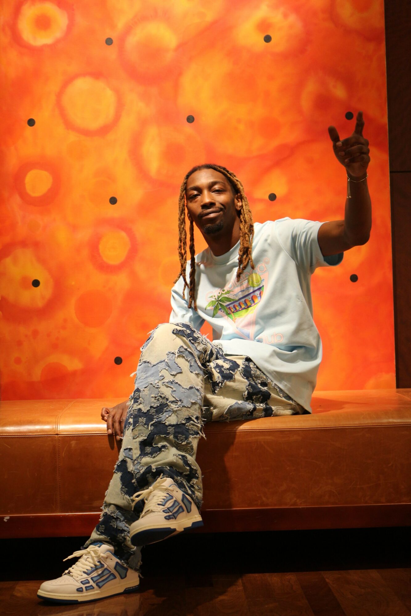 Person sitting on a bench making a peace sign with a colorful background, wearing a white t-shirt and patterned pants.