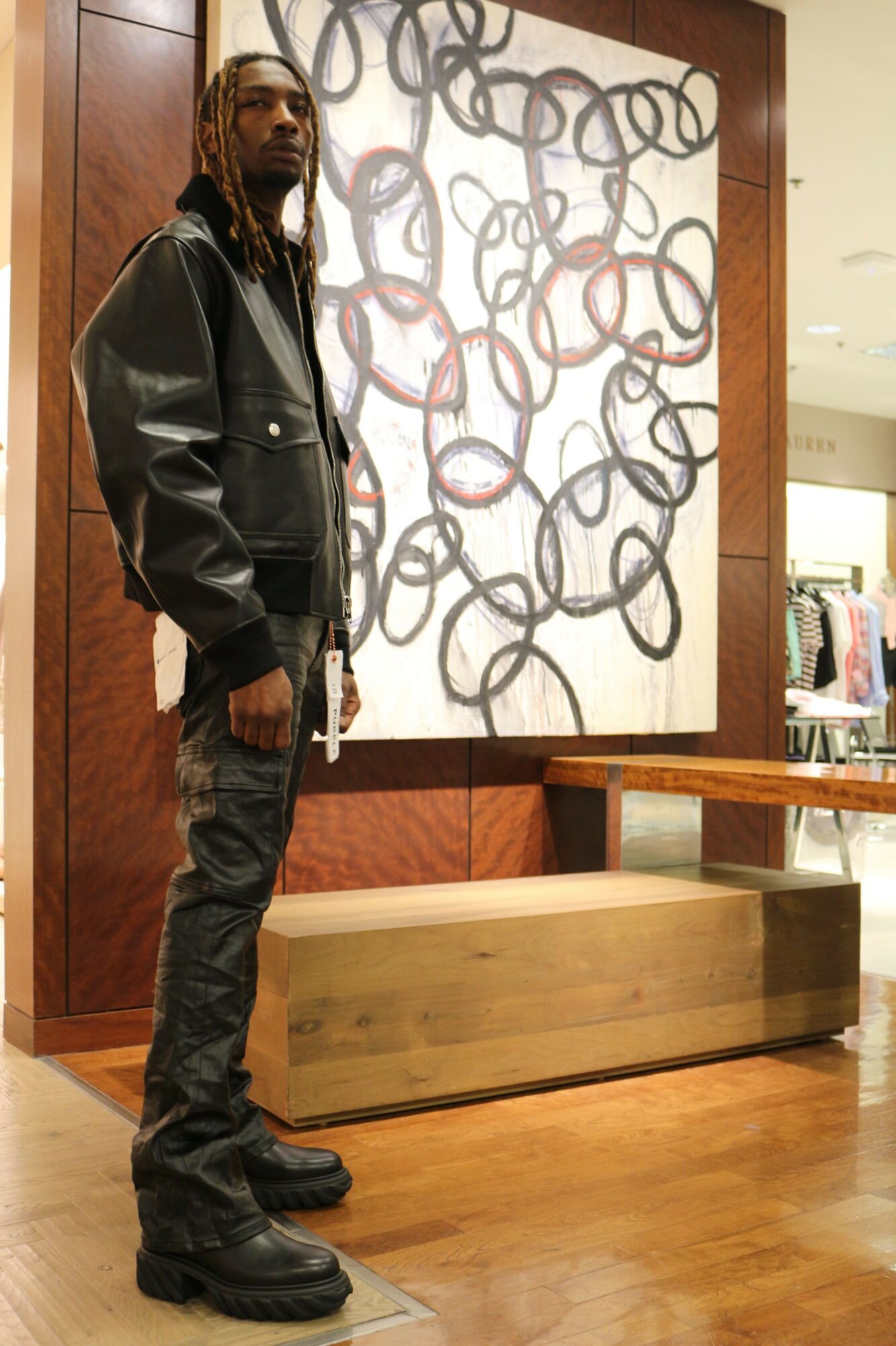 Person standing in a room with wooden flooring and furniture, wearing a black jacket, pants, and boots, facing sideways.