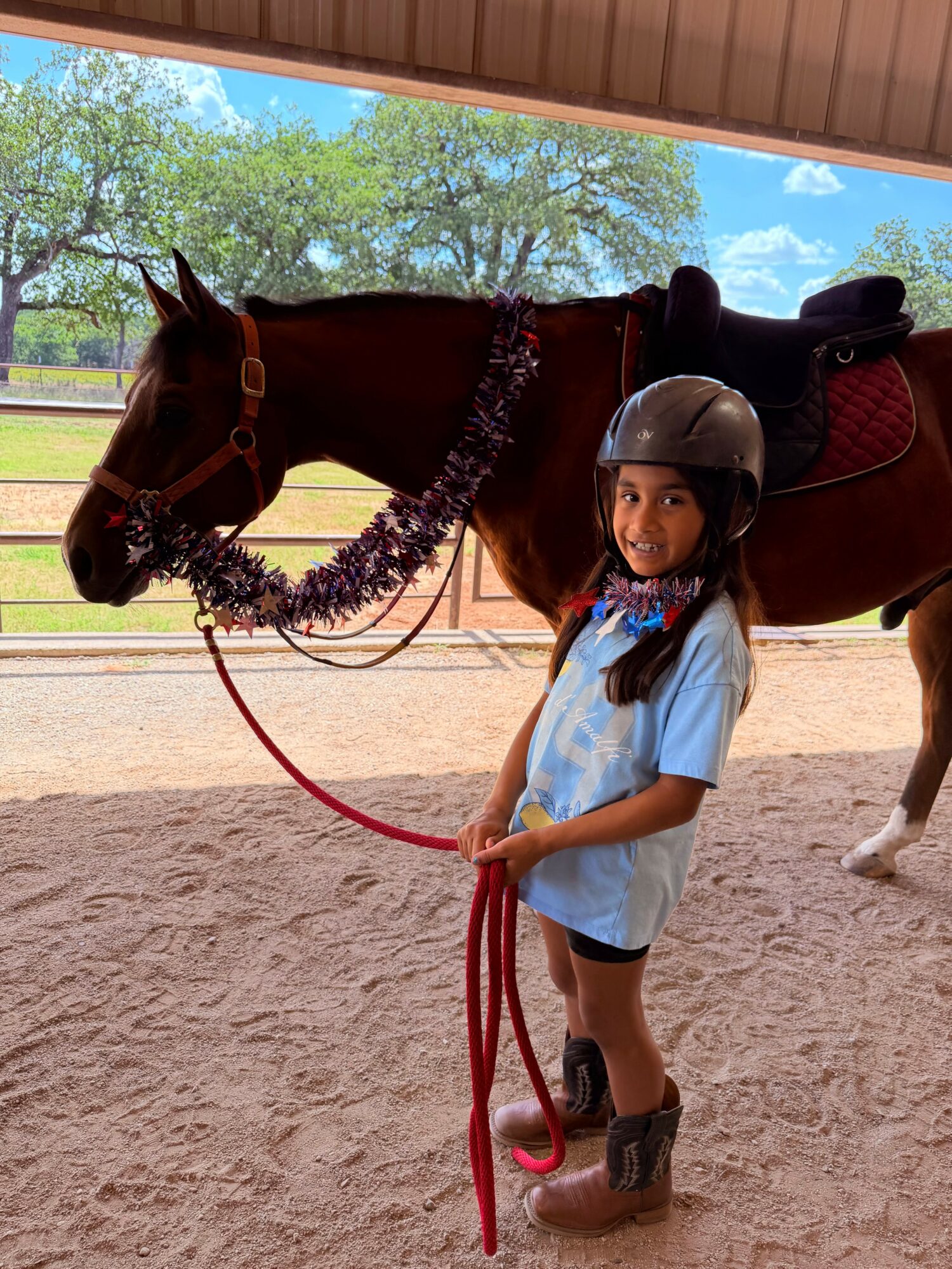 Young girl wearing a helmet and riding boots stands next to a saddled horse under a shelter, holding the horse's lead rope.