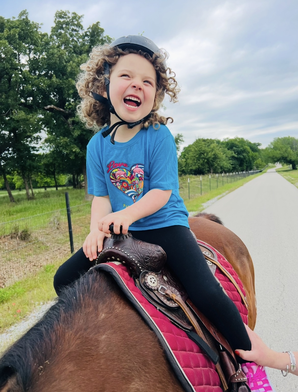 Child with curly hair smiling and laughing while riding a horse outdoors on a sunny day.