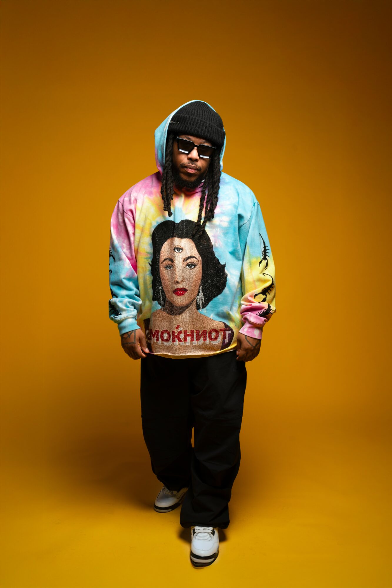 Person wearing a colorful hoodie with a woman's face and text, black pants, white shoes, and sunglasses, standing against a yellow background.