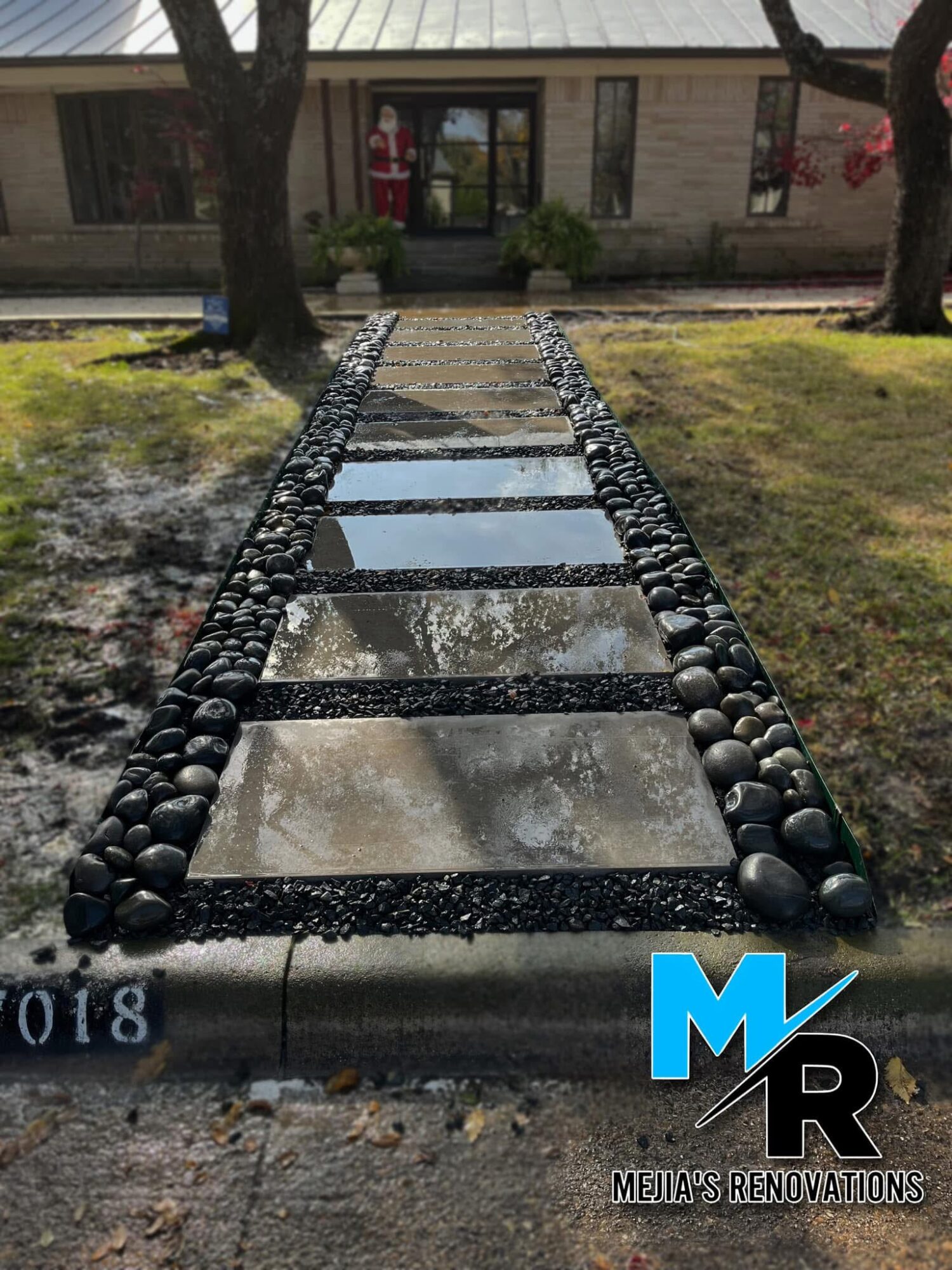 Stone pathway with rectangular tiles bordered by black pebbles, leading to a house with trees and grass on sides.