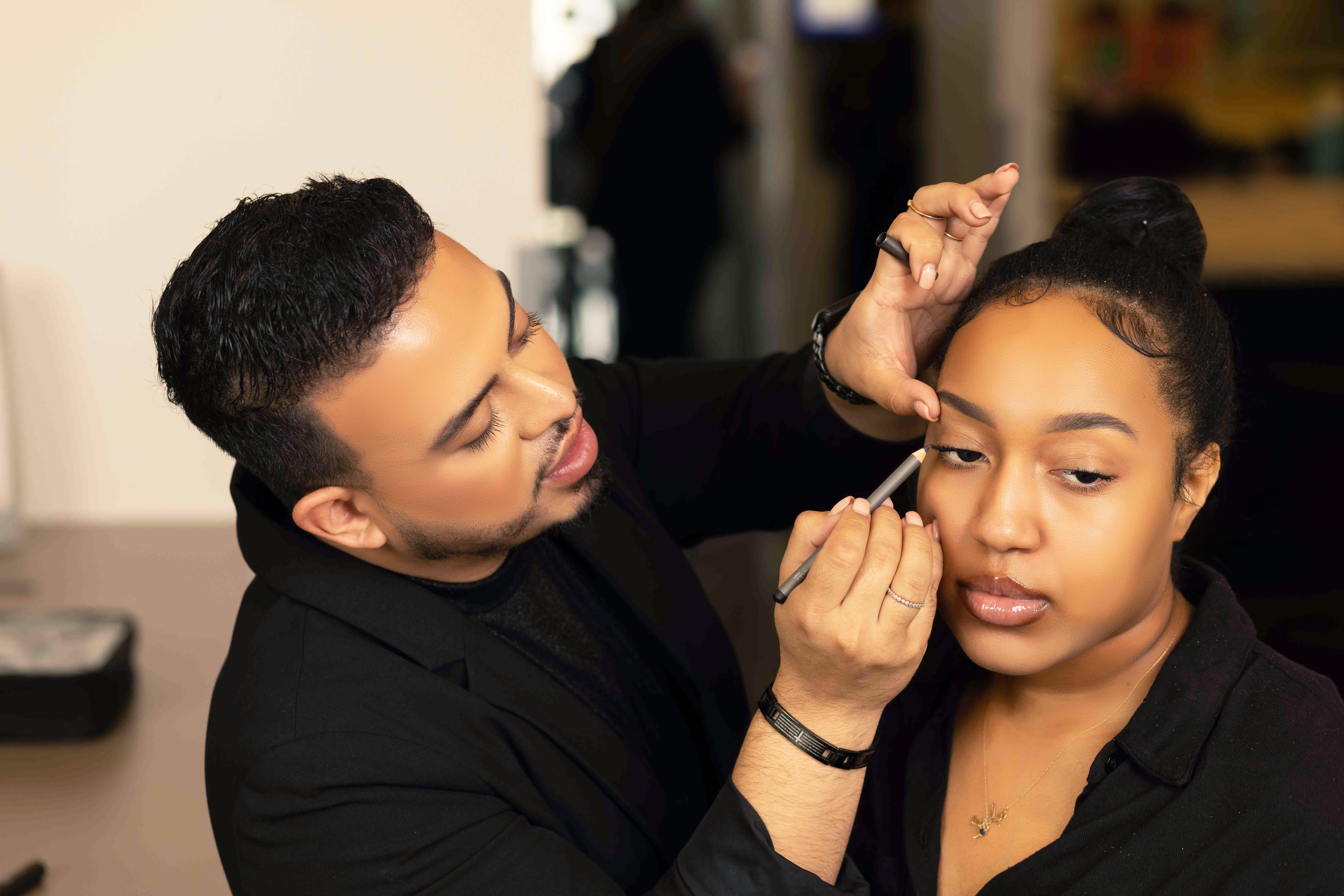 Makeup artist applies makeup to a young woman with her hair in a bun, in a well-lit room.