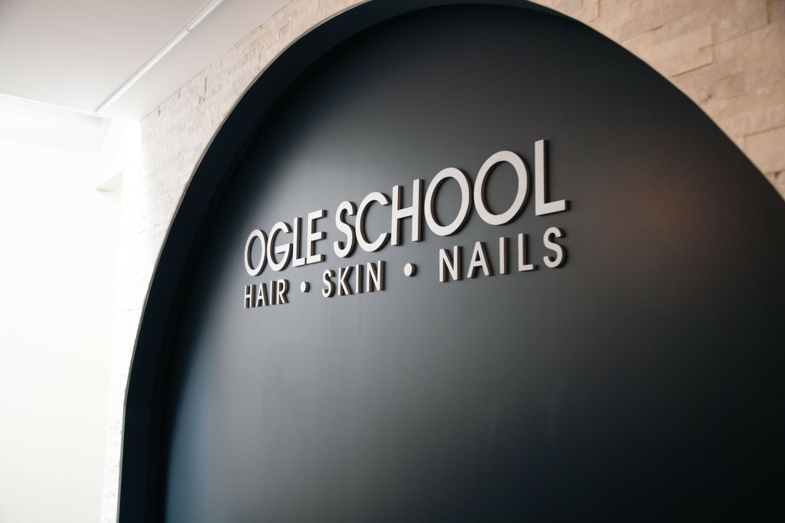Sign reading 'Ogle School' with smaller text 'Hair, Skin, Nails' on a black wall, curved background.