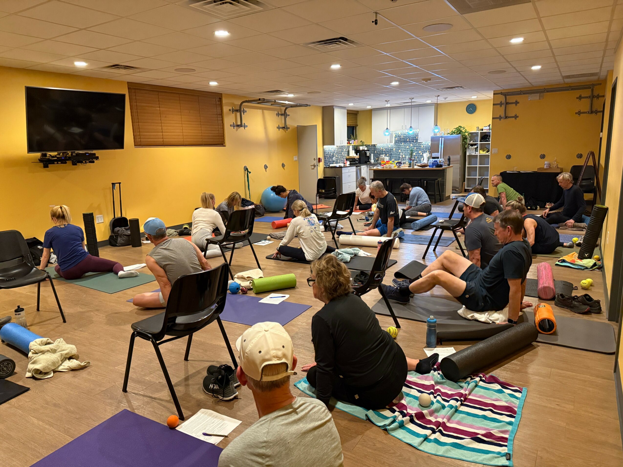 People participating in a seated yoga class in a room with yellow walls, mats, and exercise props, facing a TV screen.