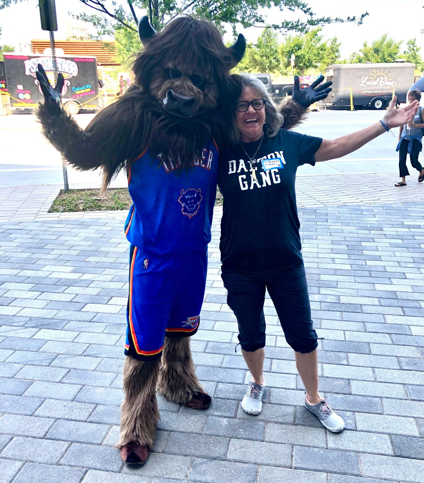 Person in a Bigfoot costume with arms raised, standing next to smiling woman with arms outstretched outdoors on paved area.
