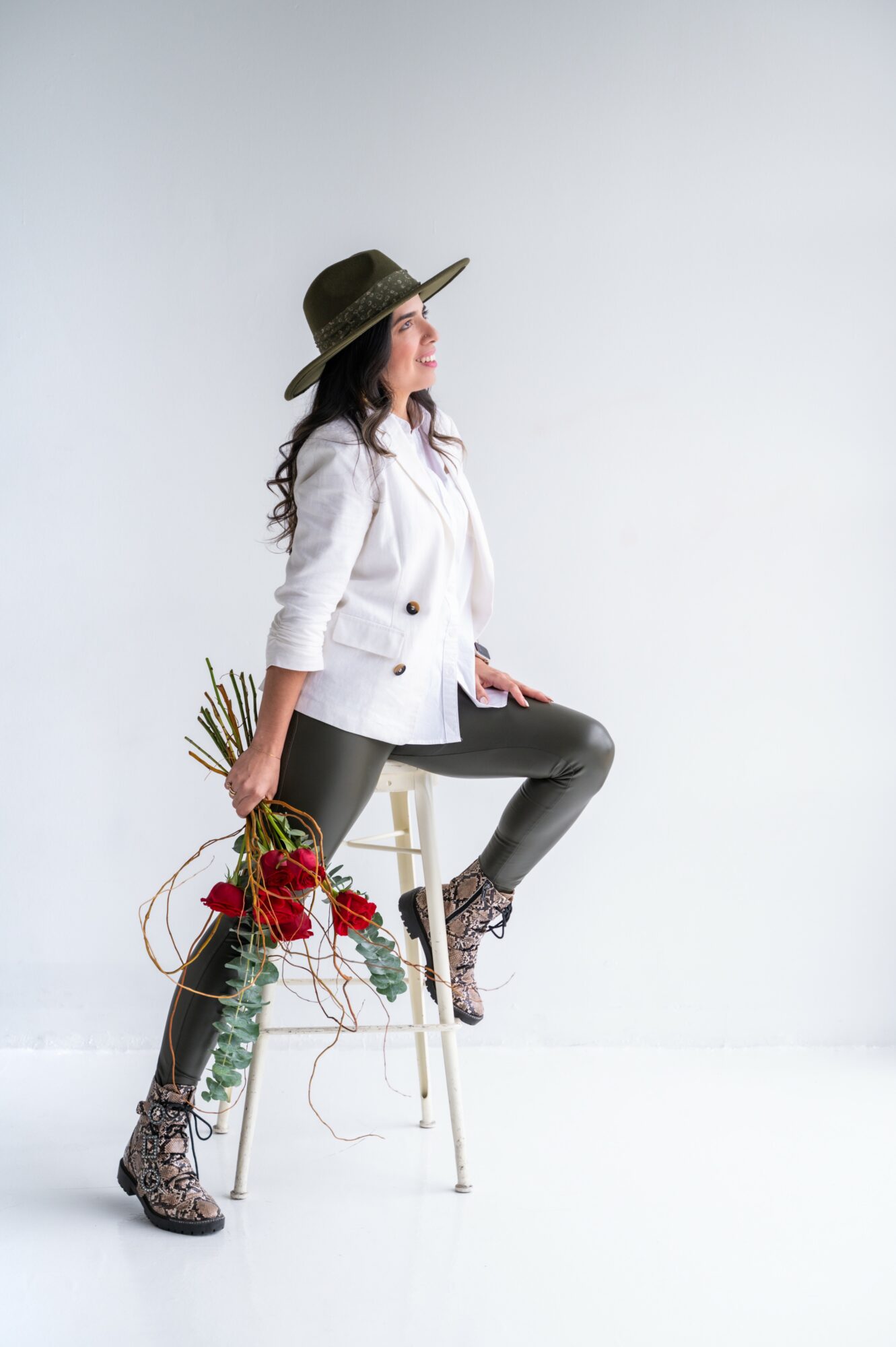 Woman in white jacket and black leather pants sitting on a stool, holding a bouquet of red roses, wearing a hat and patterned boots.
