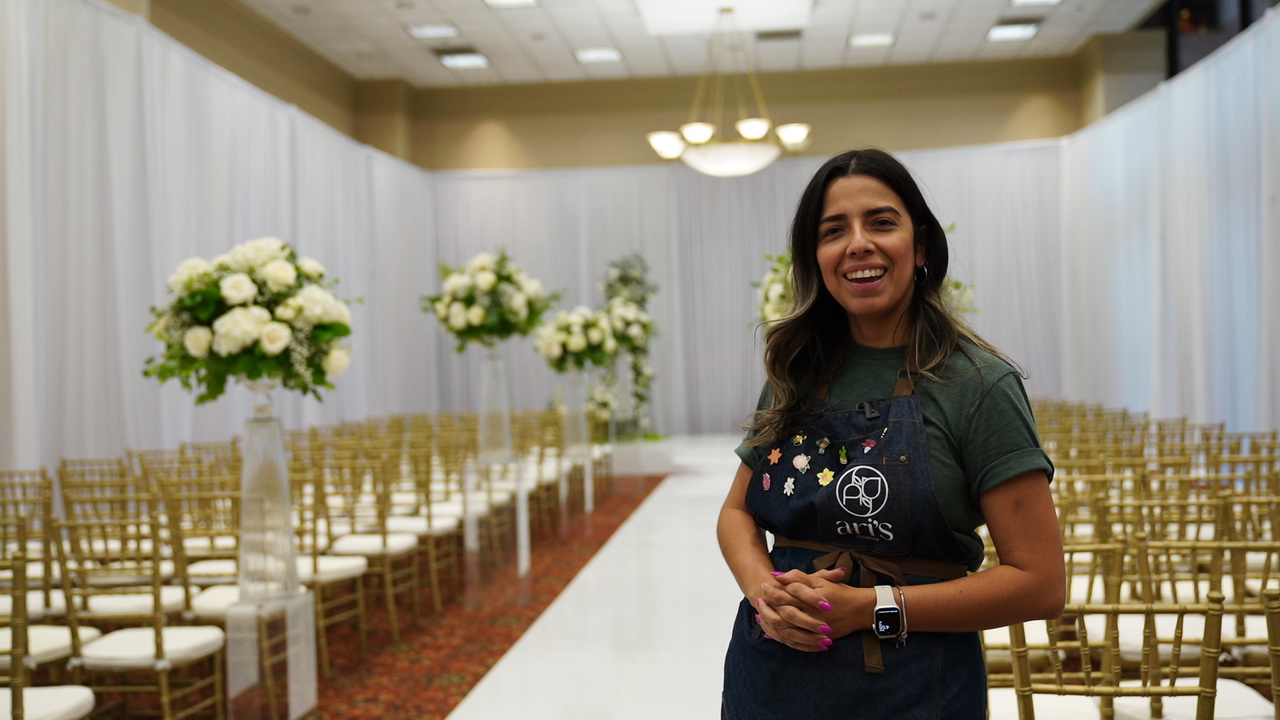 Smiling woman standing in a decorated event hall with floral arrangements and rows of chairs.