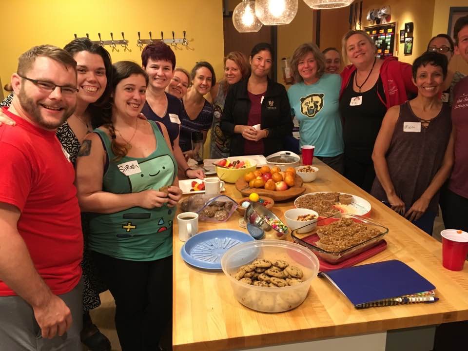 Group of people gathered around a table with food and drinks in a warmly lit room, smiling at the camera.