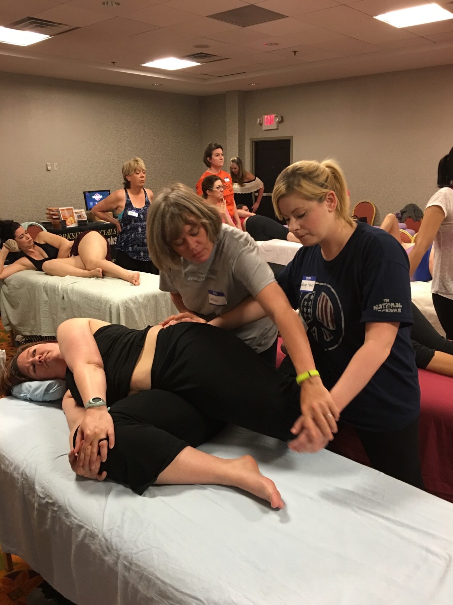 Two women assist a person lying on a bed in a room with multiple people, some sitting and some lying down, in a training or workshop setting.