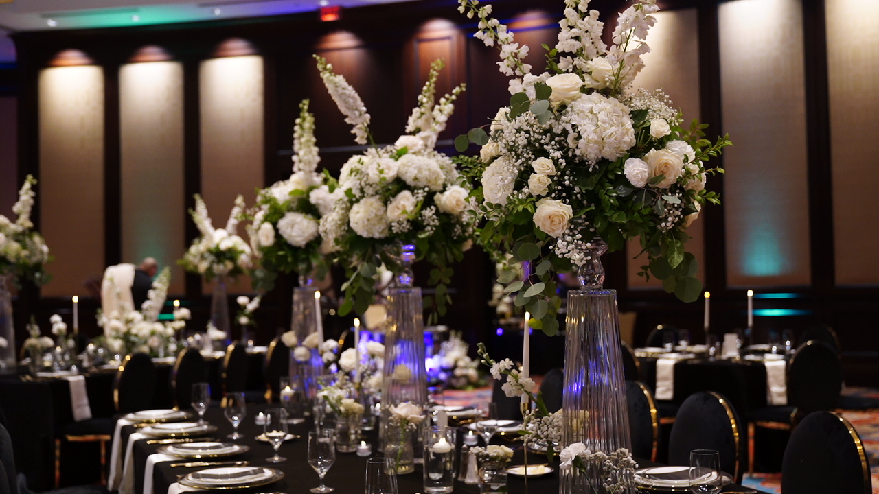 Elegant banquet table with large floral centerpieces in tall vases, set in a dimly lit event hall with colorful lighting.