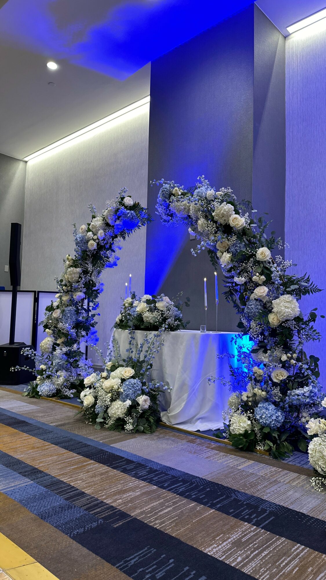 Floral arch with white and blue flowers surrounds a table with a cake, set against a modern interior backdrop.